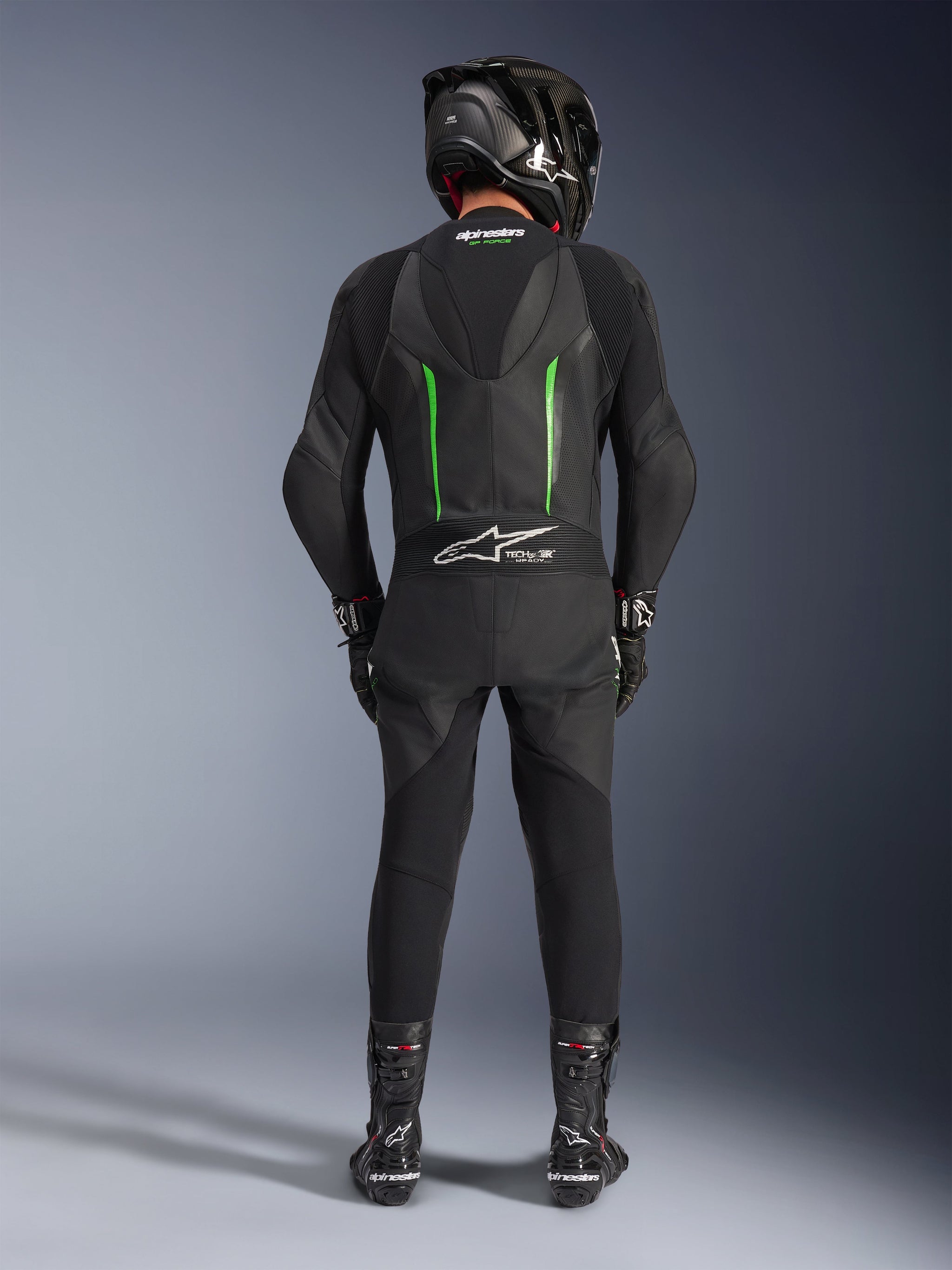 Piloto visto de trás a usar o fato de pele Alpinestars GP Force V2 - 1 Peça em preto e verde, combinado com um capacete de fibra de carbono preto, luvas de corrida pretas e botas pretas, apresentando uma corcova aerodinâmica e o logótipo Tech-Air Ready