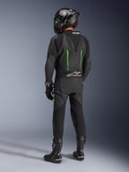 Piloto visto de costas a usar o Fato de Pele Alpinestars GP Force V2, 1 Peça, preto com detalhes em verde, corcova aerodinâmica, combinado com capacete de fibra de carbono preto, botas de corrida e luvas pretas, equipamento de motociclismo de alta performance