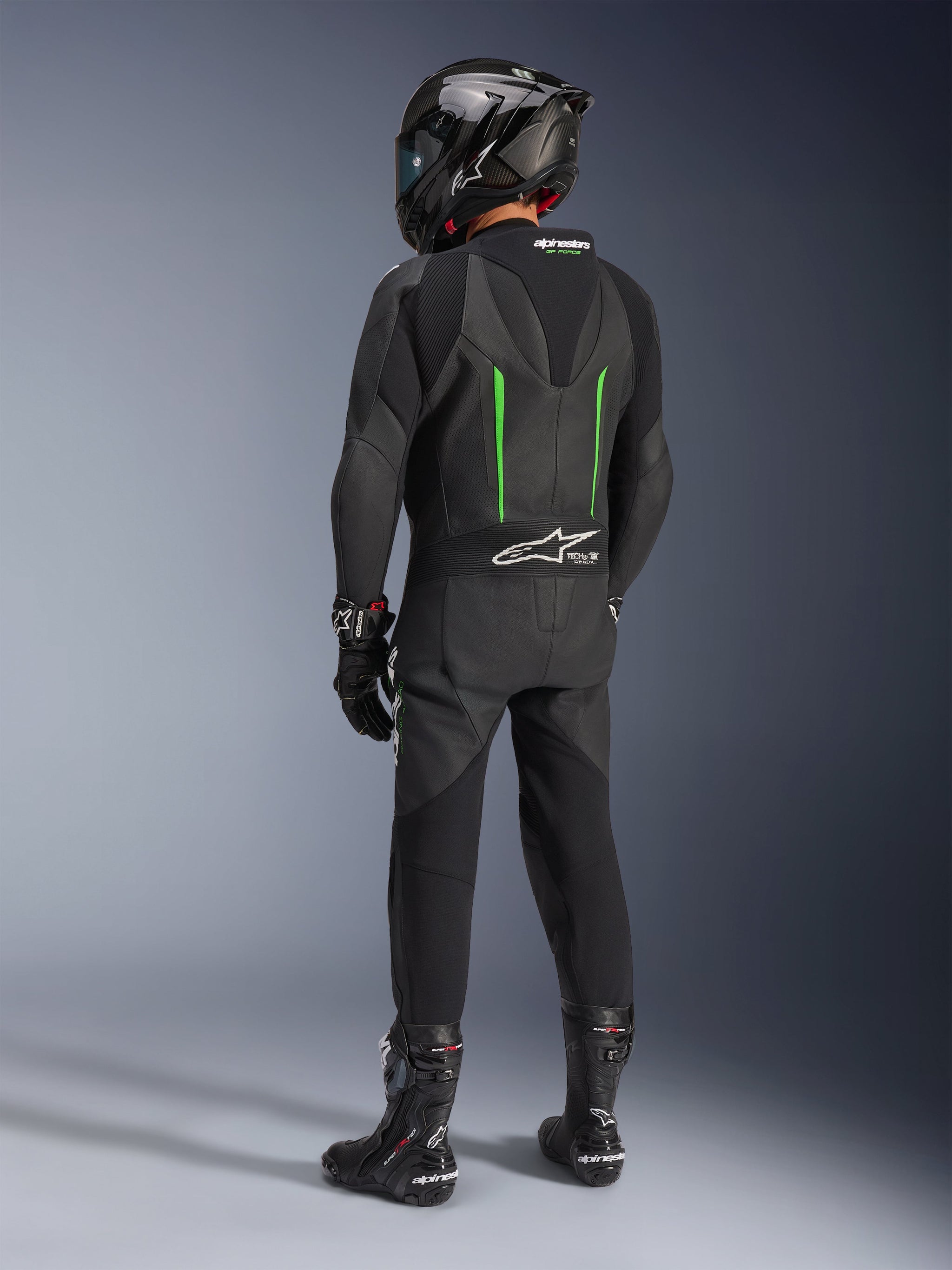 Piloto visto de costas a usar o Fato de Pele Alpinestars GP Force V2, 1 Peça, preto com detalhes em verde, corcova aerodinâmica, combinado com capacete de fibra de carbono preto, botas de corrida e luvas pretas, equipamento de motociclismo de alta performance