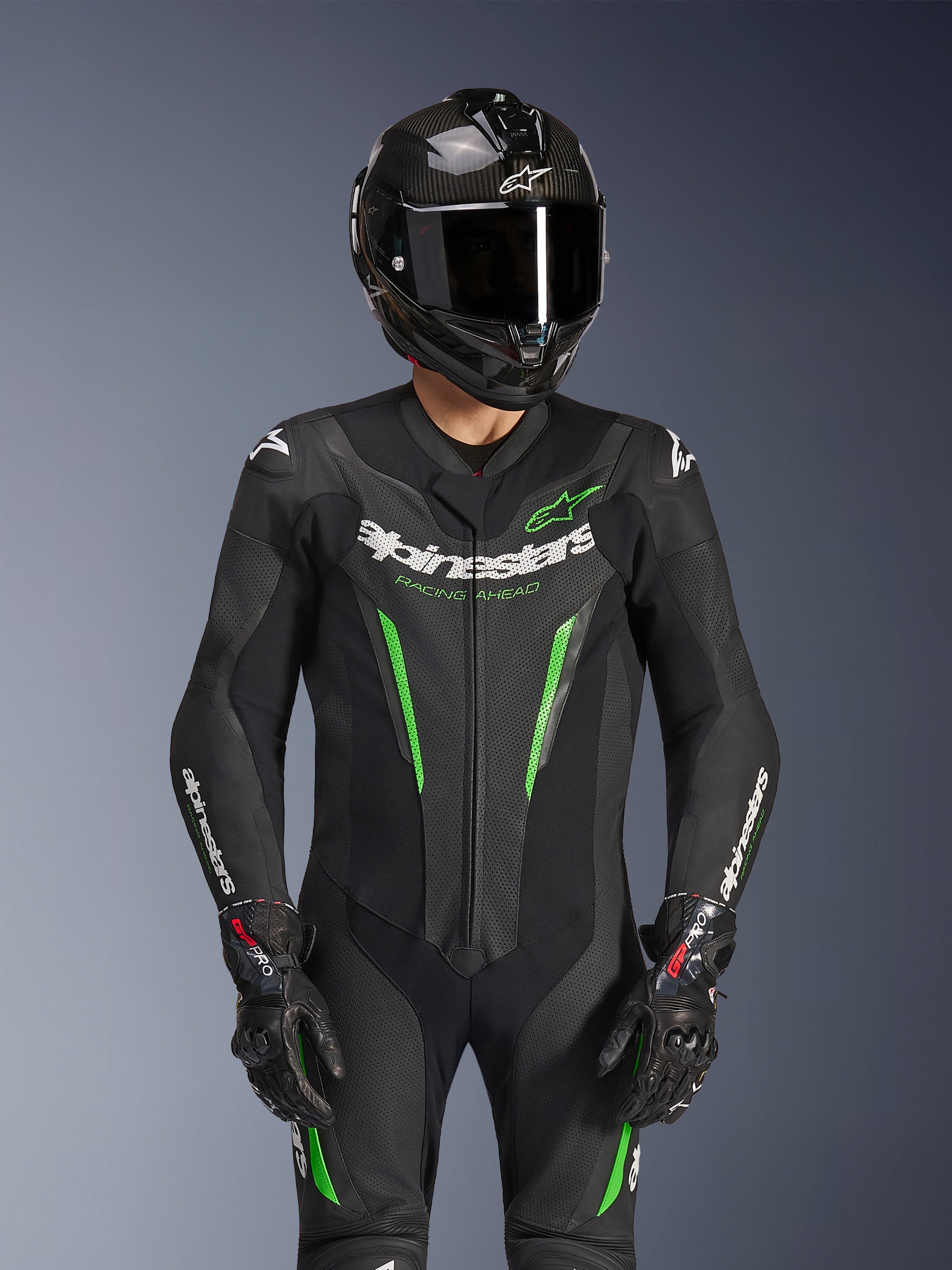 Fato de Pele Alpinestars GP Force V2 - 1 Peça, preto e verde, fato de corrida de motociclismo usado com um capacete de fibra de carbono e luvas GP Pro, com detalhes em verde lima e o logótipo Alpinestars 'Racing Ahead', concebido para o desempenho em pista.