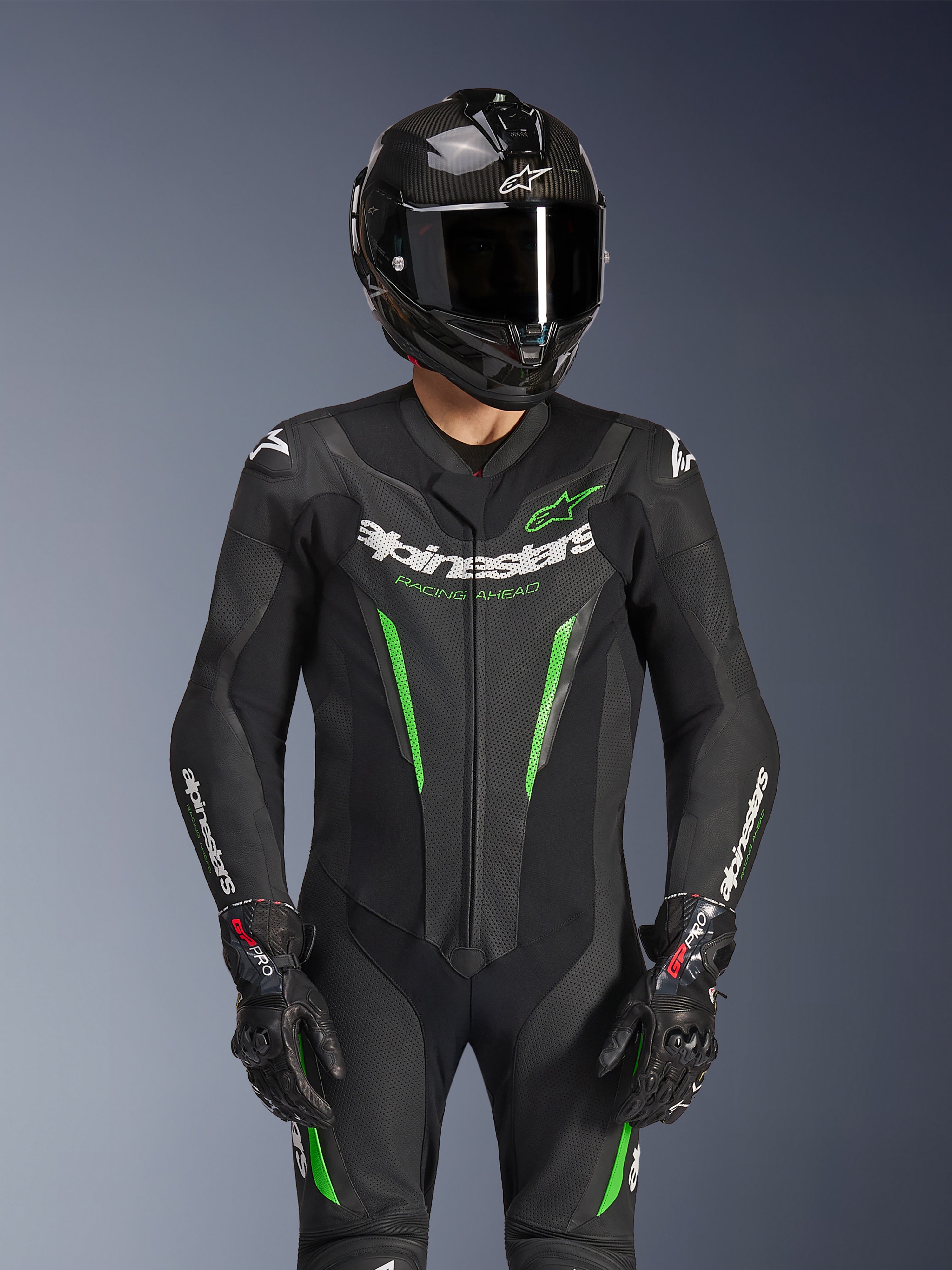 Fato de Pele Alpinestars GP Force V2 - 1 Peça, preto e verde, fato de corrida de motociclismo usado com um capacete de fibra de carbono e luvas GP Pro, com detalhes em verde lima e o logótipo Alpinestars 'Racing Ahead', concebido para o desempenho em pista.