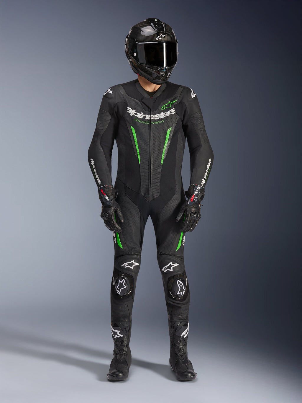 Piloto de motociclismo a usar o Fato de Pele Alpinestars GP Force V2 - 1 Peça em preto e verde, combinado com um capacete de fibra de carbono e luvas de corrida, com painéis de pele perfurada e logótipos Alpinestars brancos, equipamento profissional para pista e condução desportiva