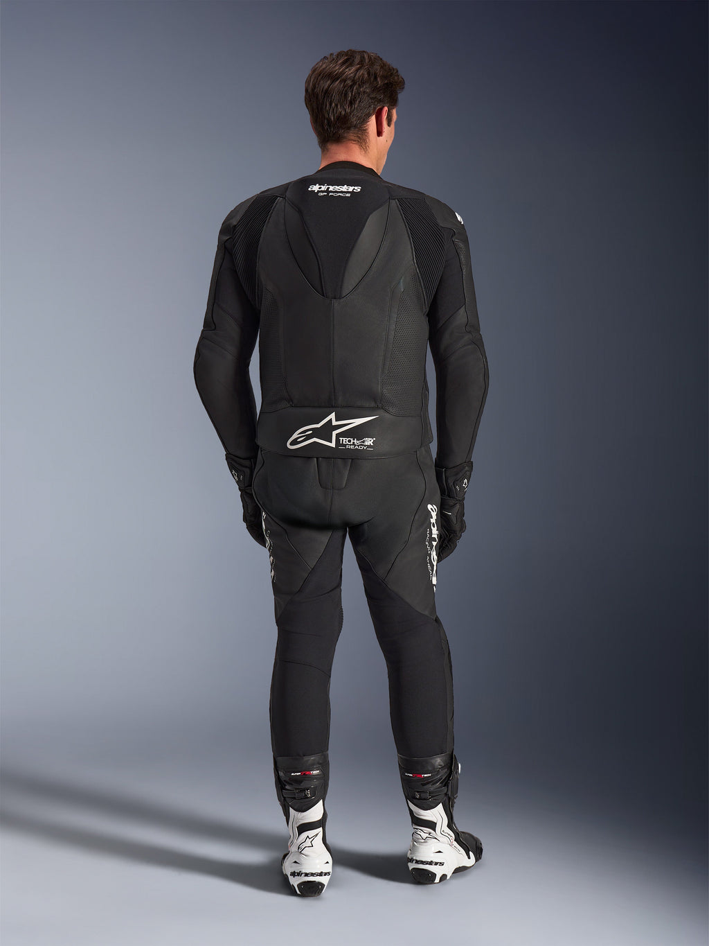 Fato de Pele Alpinestars GP Force V2 de 2 Peças, preto, vista traseira mostrando a corcova aerodinâmica e a marcação Tech-Air Ready na cintura, painéis de pele perfurada para ventilação, combinado com botas de corrida brancas e pretas, equipamento profissional de motociclismo