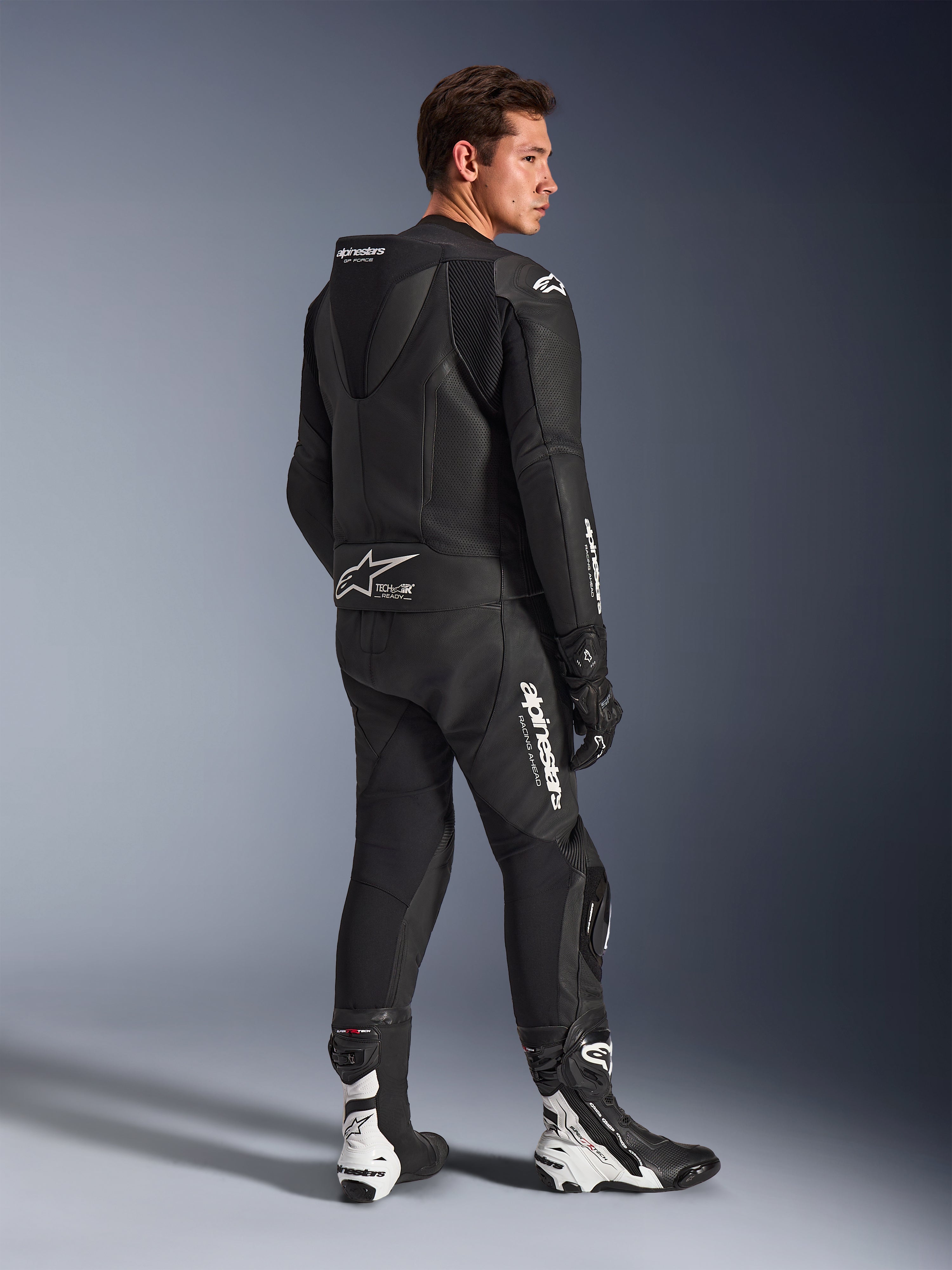 Modelo masculino a usar o Fato de Pele Alpinestars GP Force V2 de 2 Peças em preto com logótipos brancos, vista traseira de três quartos mostrando a corcova aerodinâmica e a marca Tech-Air Ready, combinado com botas e luvas de corrida pretas e brancas, equipamento profissional de motociclismo para desporto e pista.