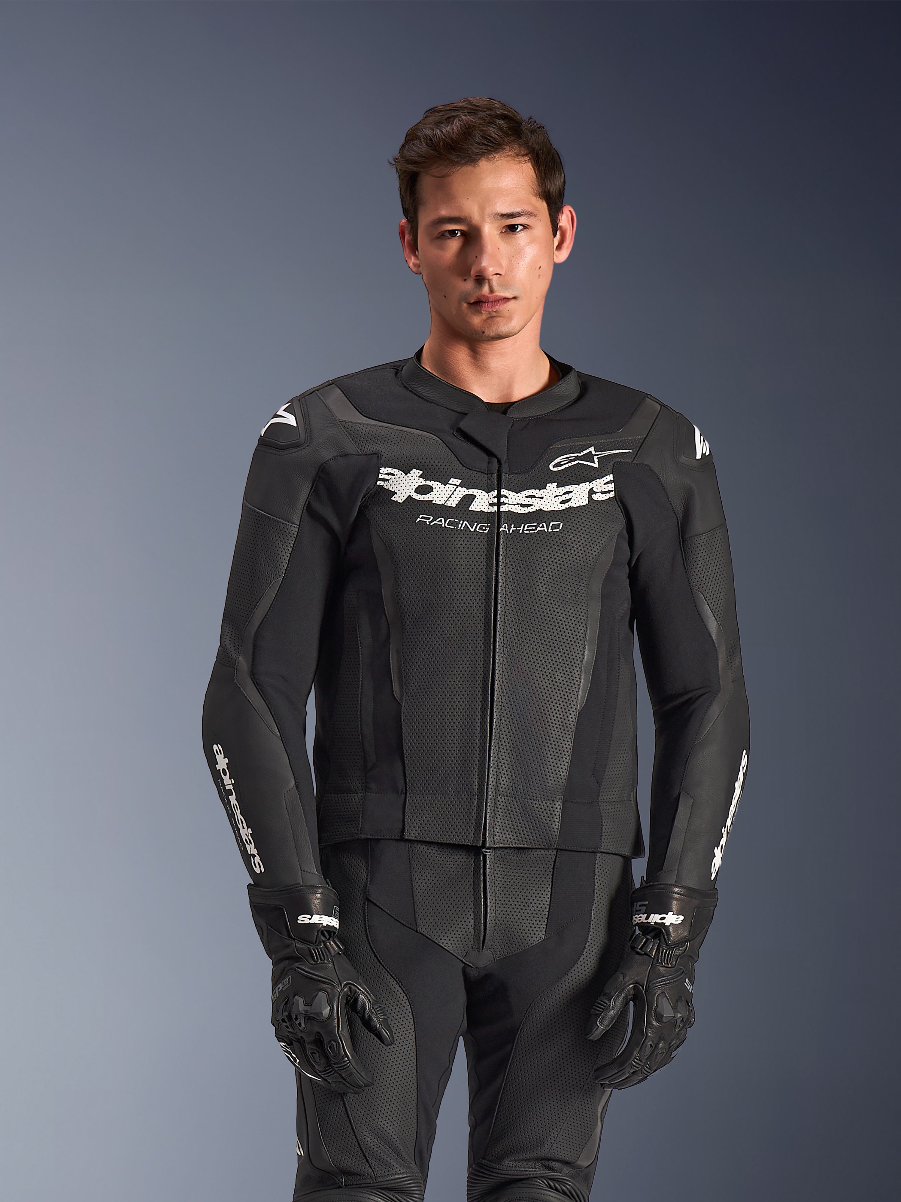 Fato de Cabedal Alpinestars GP Force V2 de 2 Peças, preto, usado por um modelo, com painéis de cabedal perfurado e logótipo branco 'Alpinestars Racing Ahead' no peito, combinado com luvas pretas, equipamento profissional de condução desportiva