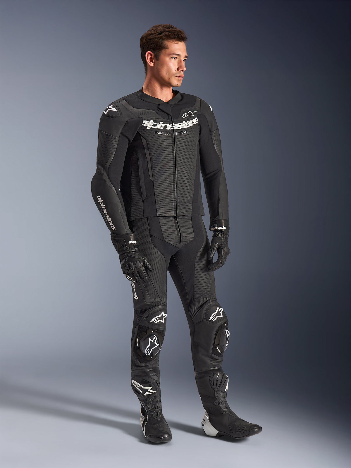 Fato de Pele Alpinestars GP Force V2 de 2 Peças, preto, usado por um modelo masculino com luvas e botas de corrida pretas a combinar, com logótipos Alpinestars em branco, painéis de pele perfurada para ventilação e sliders de joelho integrados, equipamento profissional de motociclismo