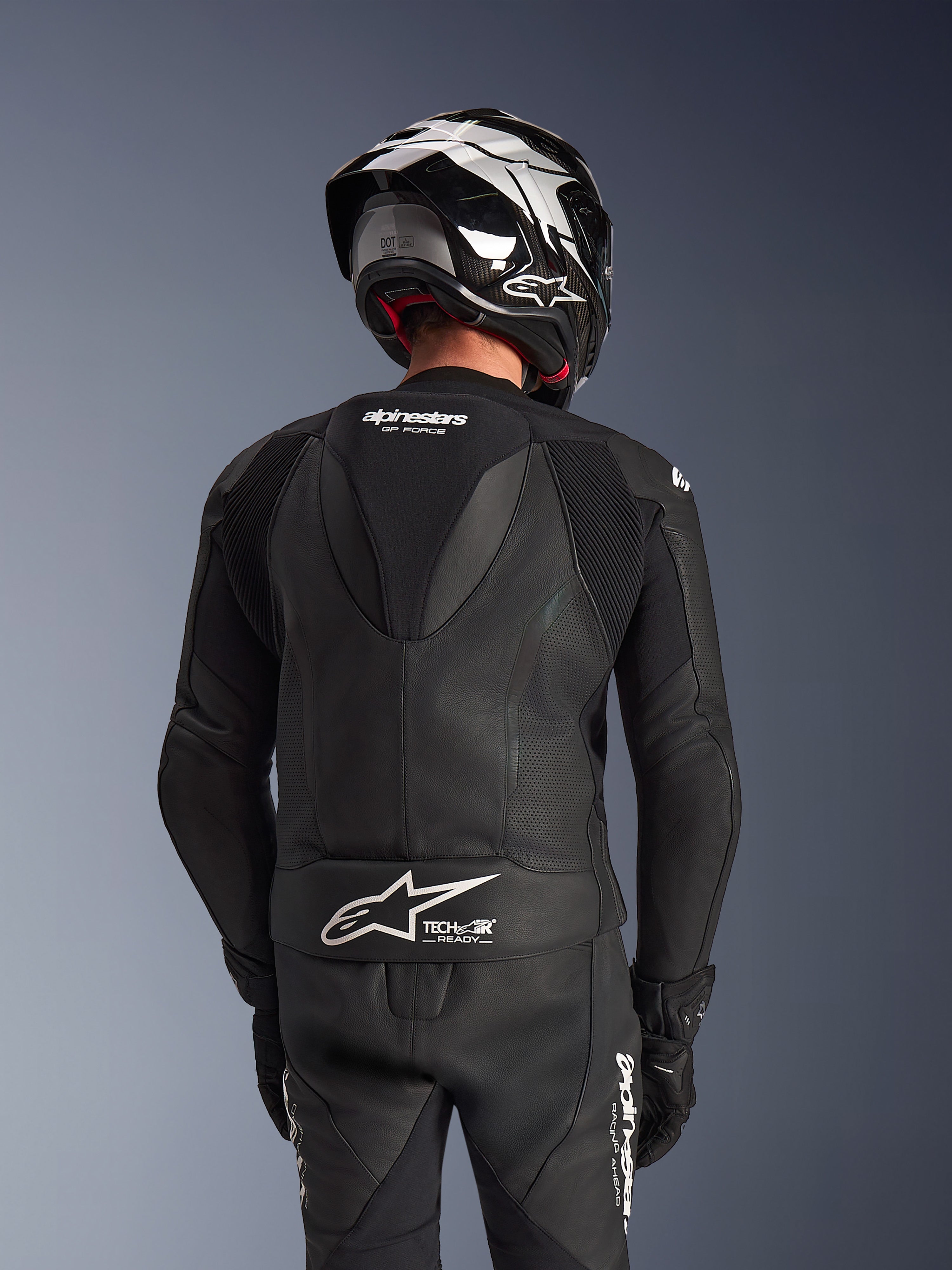 Fato de pele Alpinestars GP Force V2 de 2 peças, preto, vista traseira do piloto mostrando a corcova aerodinâmica, painéis de pele perfurada e zonas elásticas em acordeão nos ombros, com marca Tech-Air Ready na parte inferior das costas, usado com capacete de corrida e luvas a condizer.