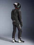 Alpinestars GP Force V2 2-Piece Leather Suit, preto, vista traseira de motociclista com equipamento de corrida completo, com corcova aerodinâmica, logótipos brancos e pele perfurada, combinado com um capacete de fibra de carbono e botas de corrida pretas e brancas