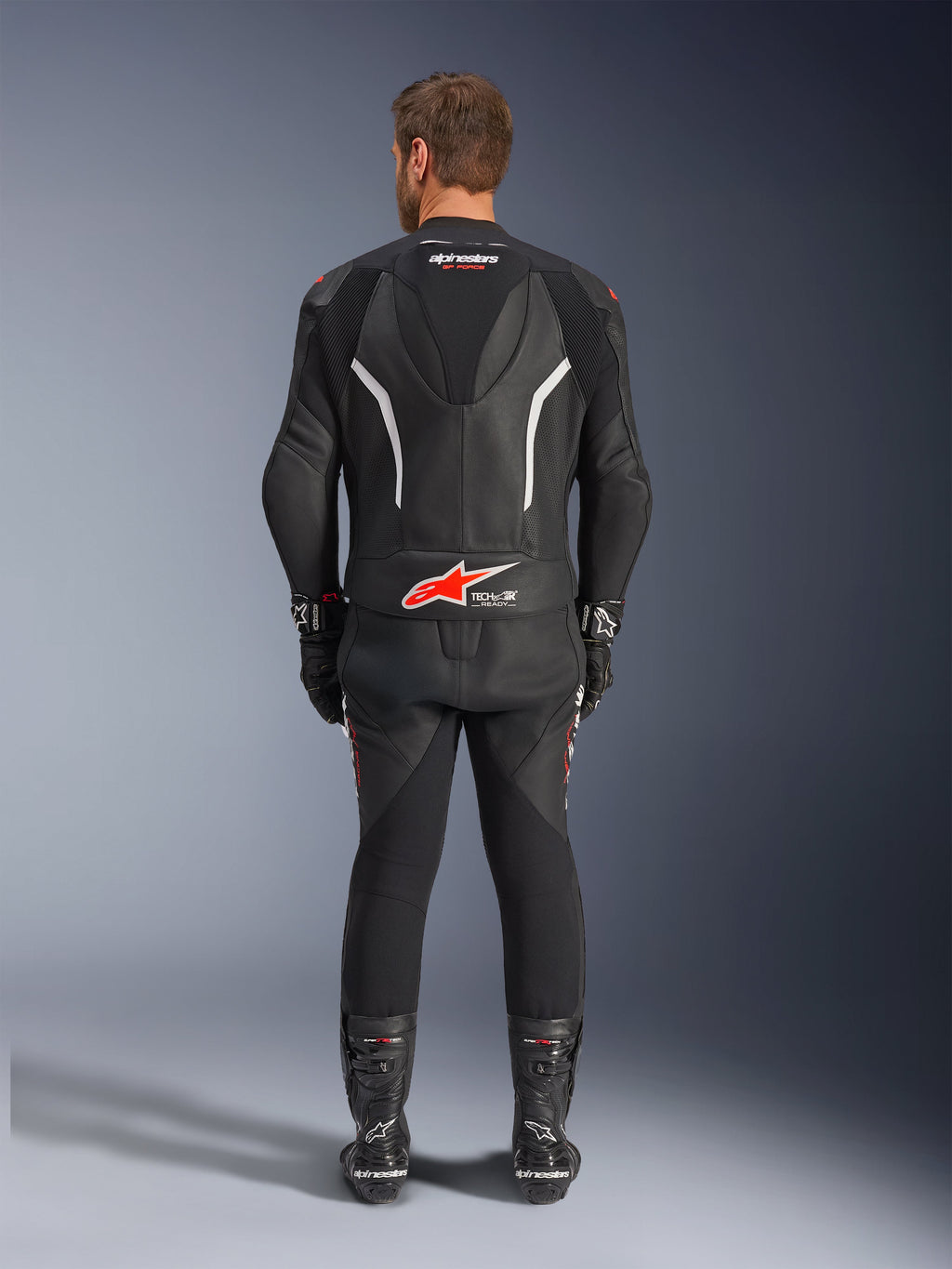 Vista traseira de um piloto a usar o Fato de Pele de 2 Peças Alpinestars GP Force V2, preto com detalhes em branco e vermelho, apresentando as marcas Alpinestars e Tech-Air Ready em destaque nas costas, complementado com luvas e botas de corrida pretas a condizer.