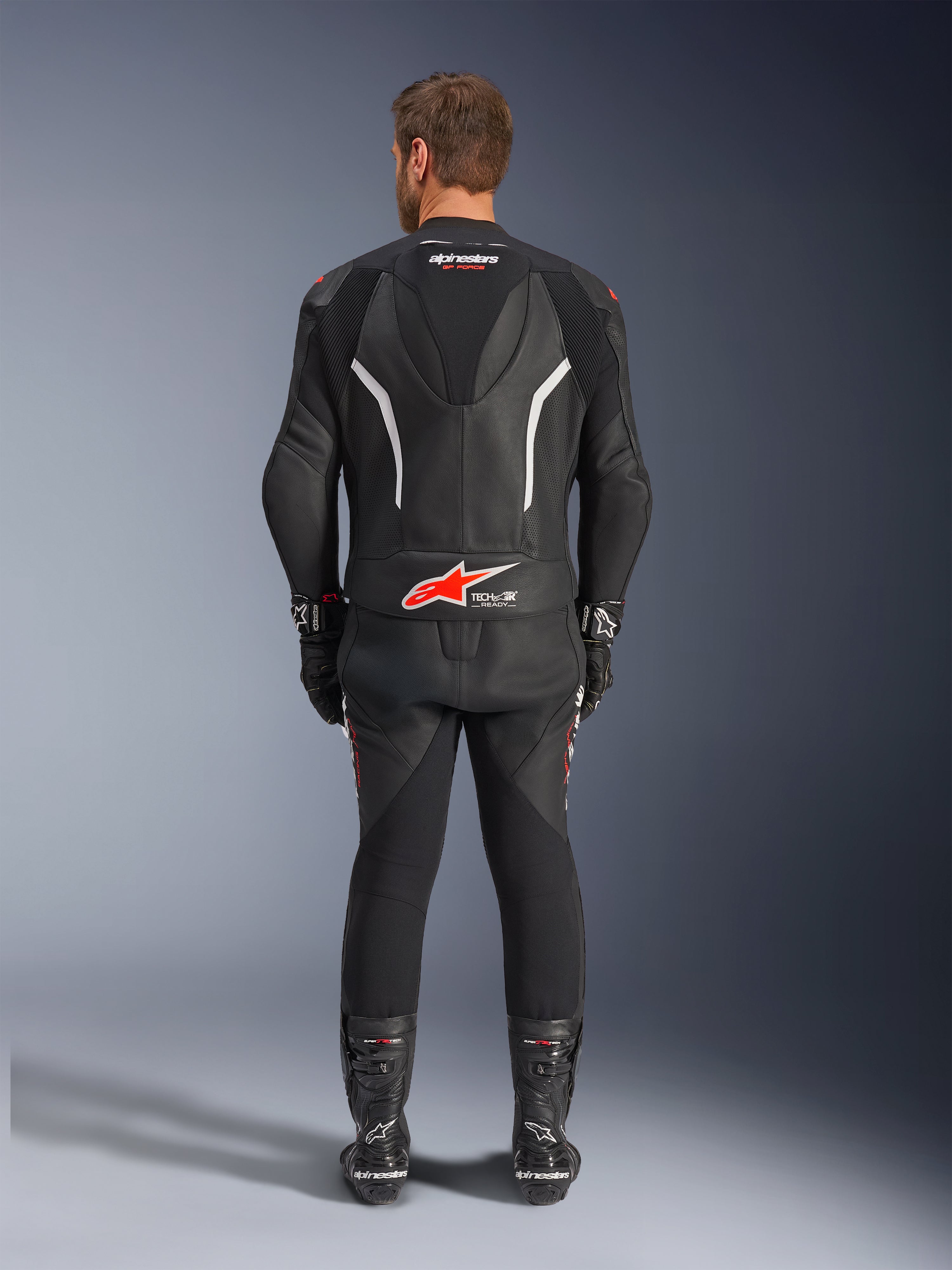 Vista traseira de um piloto a usar o Fato de Pele de 2 Peças Alpinestars GP Force V2, preto com detalhes em branco e vermelho, apresentando as marcas Alpinestars e Tech-Air Ready em destaque nas costas, complementado com luvas e botas de corrida pretas a condizer.