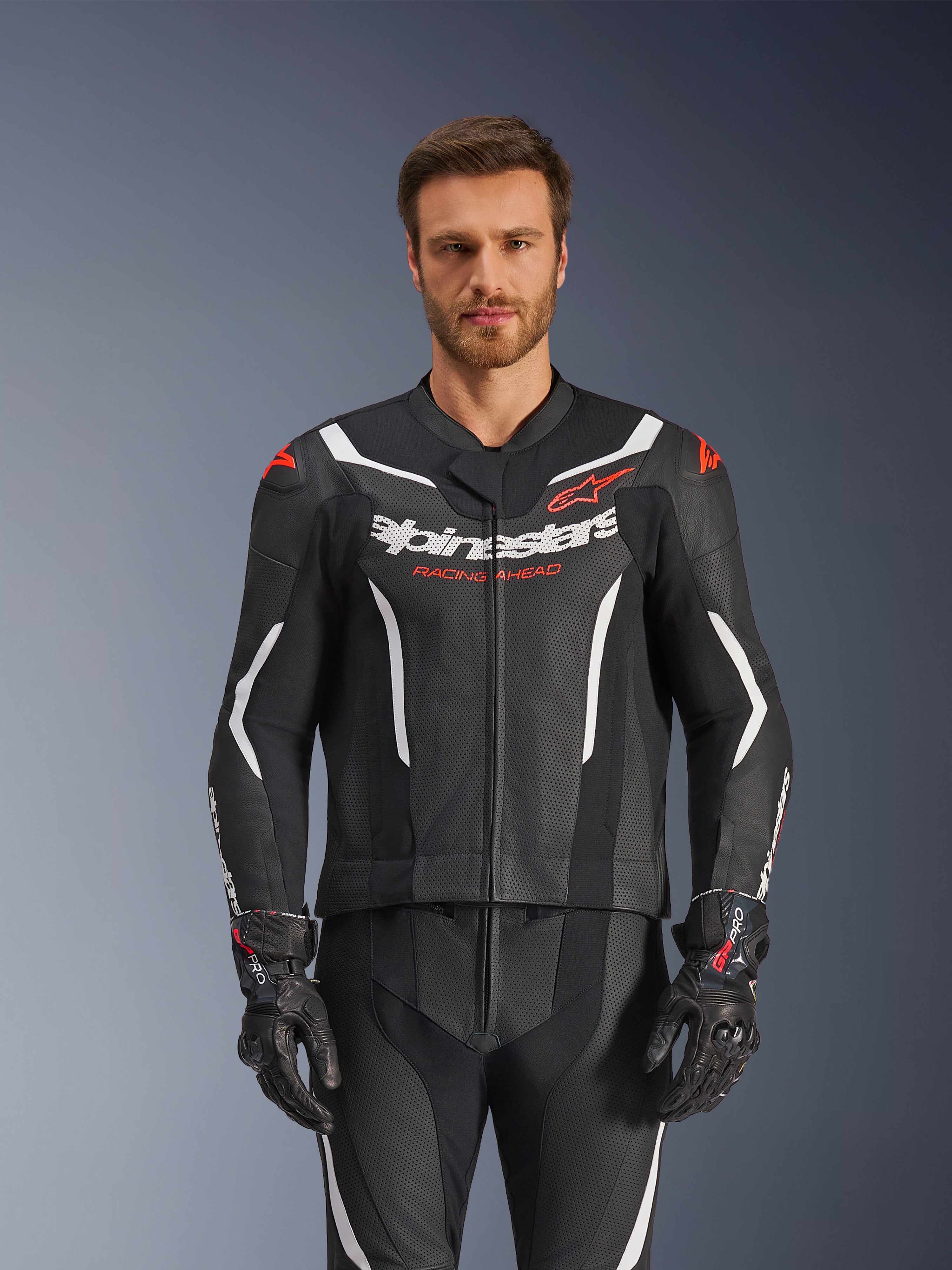 Alpinestars GP Force V2 Leather Suit - 2 Peças, preto, branco e vermelho fluo, vista frontal de modelo masculino a usar o fato de pele perfurada com luvas pretas a condizer, apresentando detalhes angulares brancos e a marca Alpinestars no peito