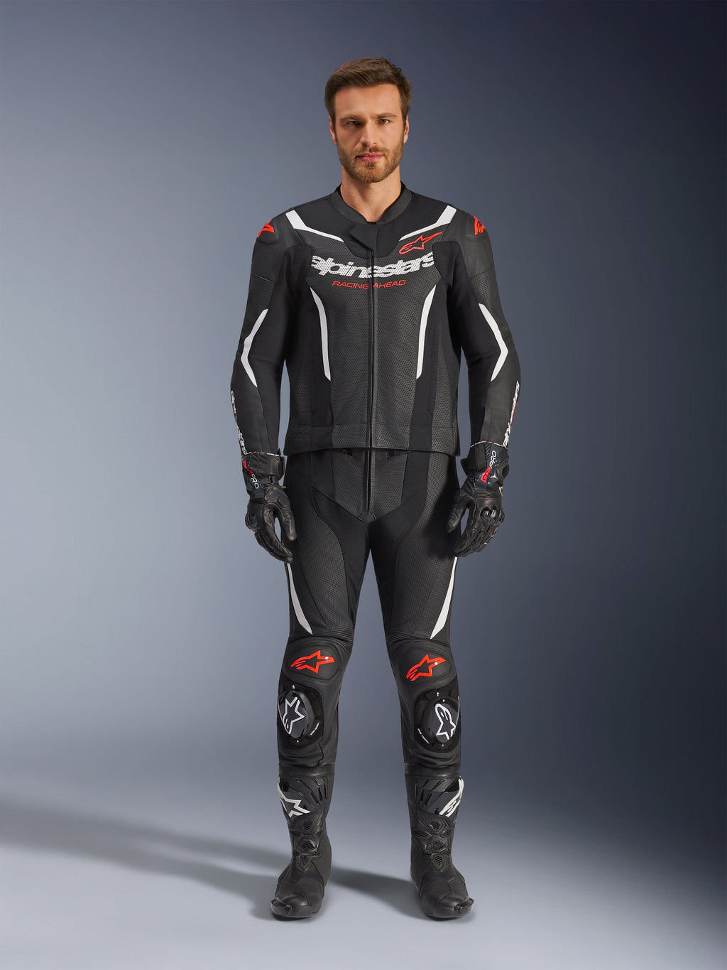 Alpinestars GP Force V2 Leather Suit de 2 Peças, fato de motociclismo de corrida, preto, branco e vermelho fluo, vestido por um modelo com luvas e botas pretas a condizer, com painéis de pele perfurada, detalhes em branco e vermelho fluo e um design aerodinâmico elegante para pista e condução desportiva