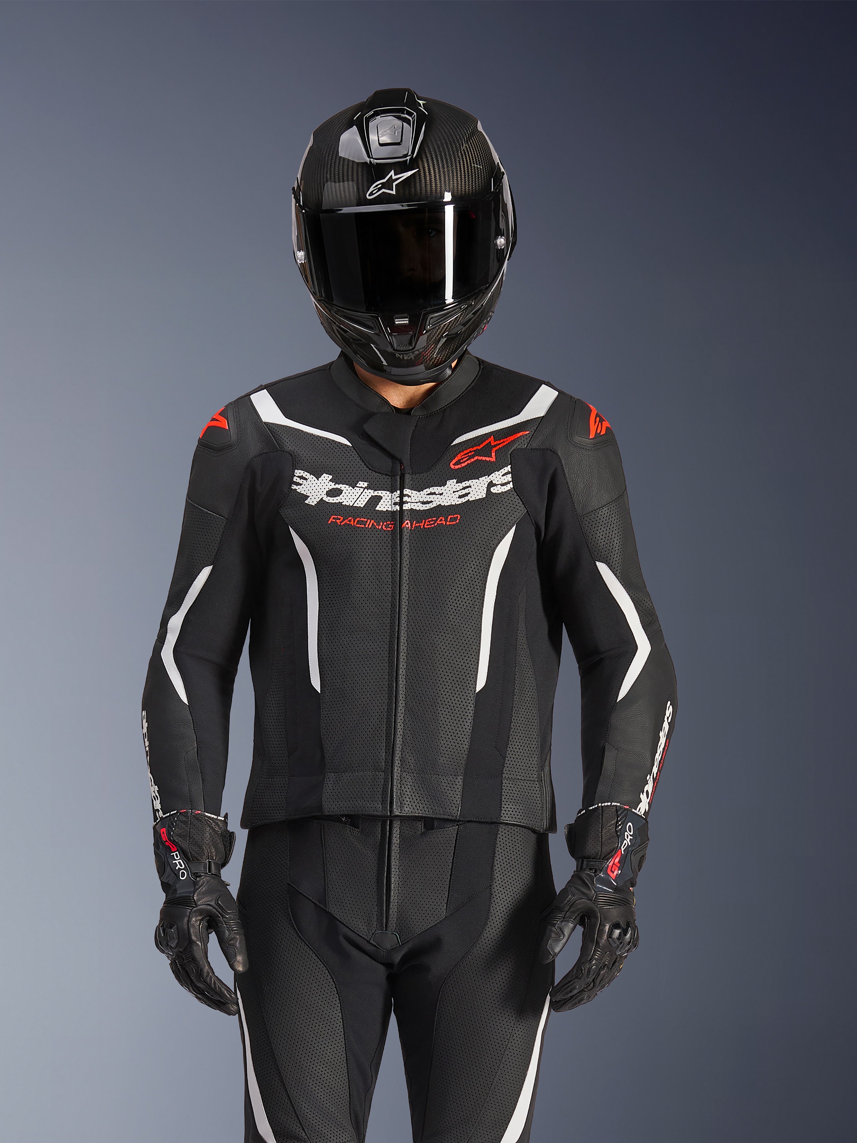 Fato de Pele Alpinestars GP Force V2 de 2 Peças, preto, branco e vermelho fluo, usado por um piloto com capacete integral de fibra de carbono e luvas de corrida pretas, com painéis de pele perfurada e logótipo Alpinestars no peito