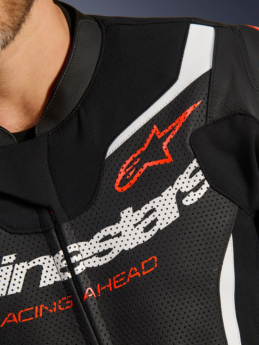 Alpinestars GP Force V2 Leather Suit 2-Piece, grande plano do peito, pele perfurada preta com detalhes em branco e vermelho, destacando o logótipo Alpinestars vermelho e o texto 'RACING AHEAD', detalhe de vestuário de competição de motociclismo premium