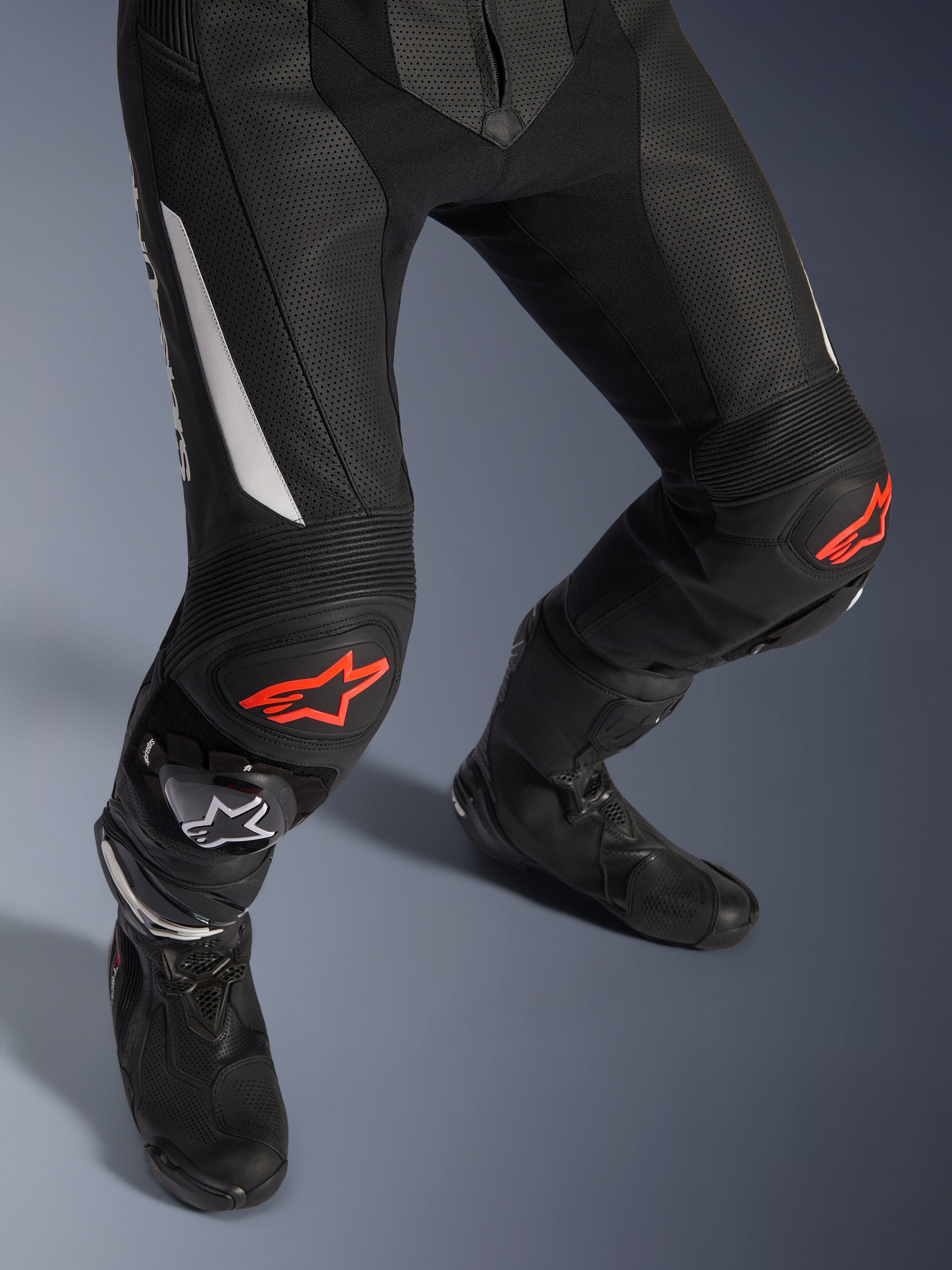 Fato de Pele Alpinestars GP Force V2 de 2 Peças, calças de motociclismo em preto com detalhes em vermelho e branco, com pele perfurada, painéis extensíveis em acordeão acima dos joelhos com logótipos de estrela vermelhos, usadas com botas de motociclismo pretas, equipamento de corrida de alta performance