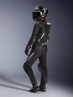 Motociclista feminina a usar o Fato de Cabedal de 2 Peças Alpinestars Stella GP-R7 em Preto, capacete de corrida integral com acabamento em carbono, luvas e botas a condizer, vista traseira realçando a corcova aerodinâmica e os logótipos brancos da Alpinestars, equipamento profissional de pista e condução desportiva