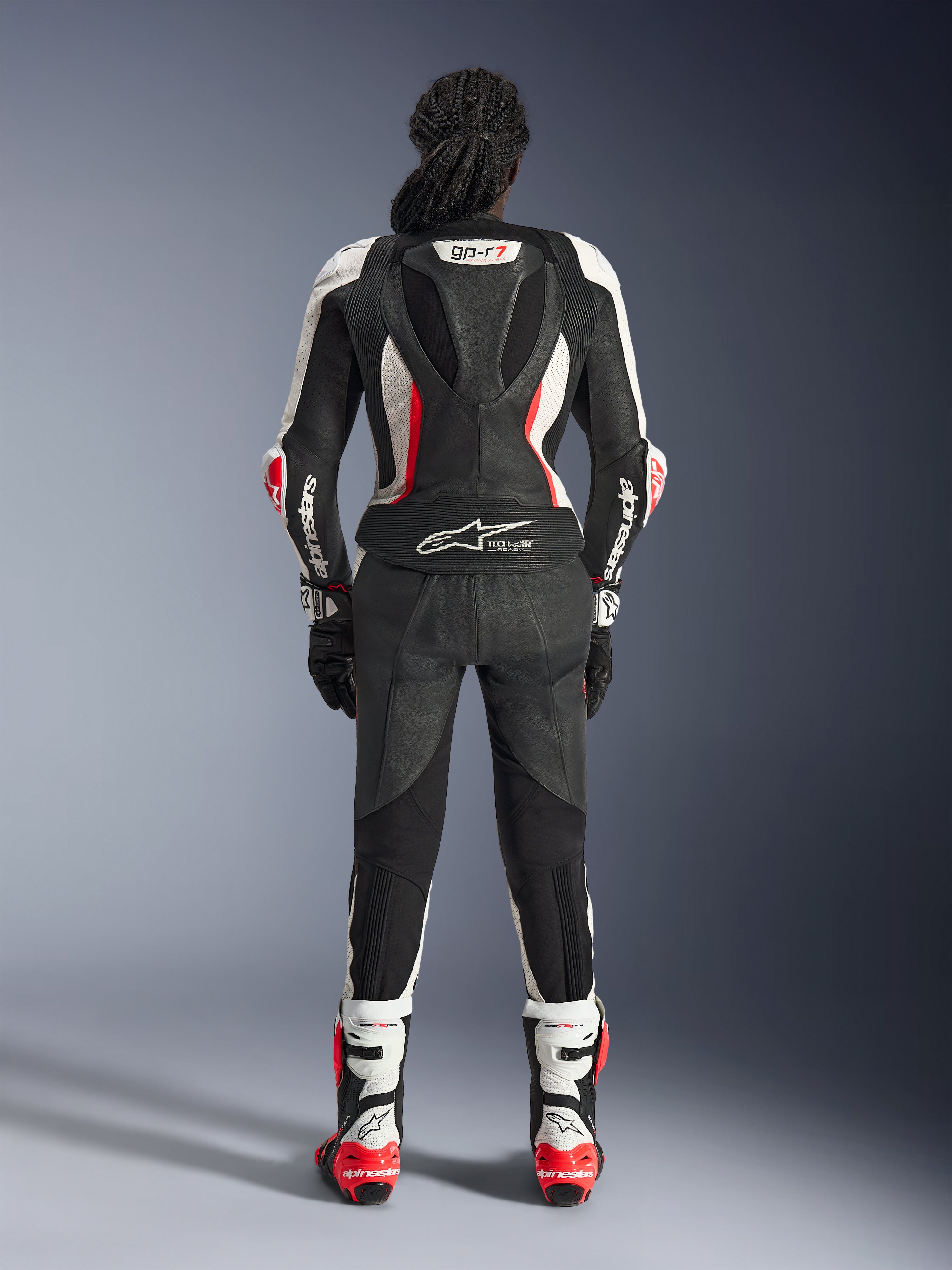 Rear view of the Alpinestars Stella GP-R7 2-Piece Leather Suit, Black White and Red Fluo, featuring an aerodynamic hump with GP-R7 branding and Tech-Air Ready logo, worn with matching racing boots and gloves, professional motorcycle gear for women