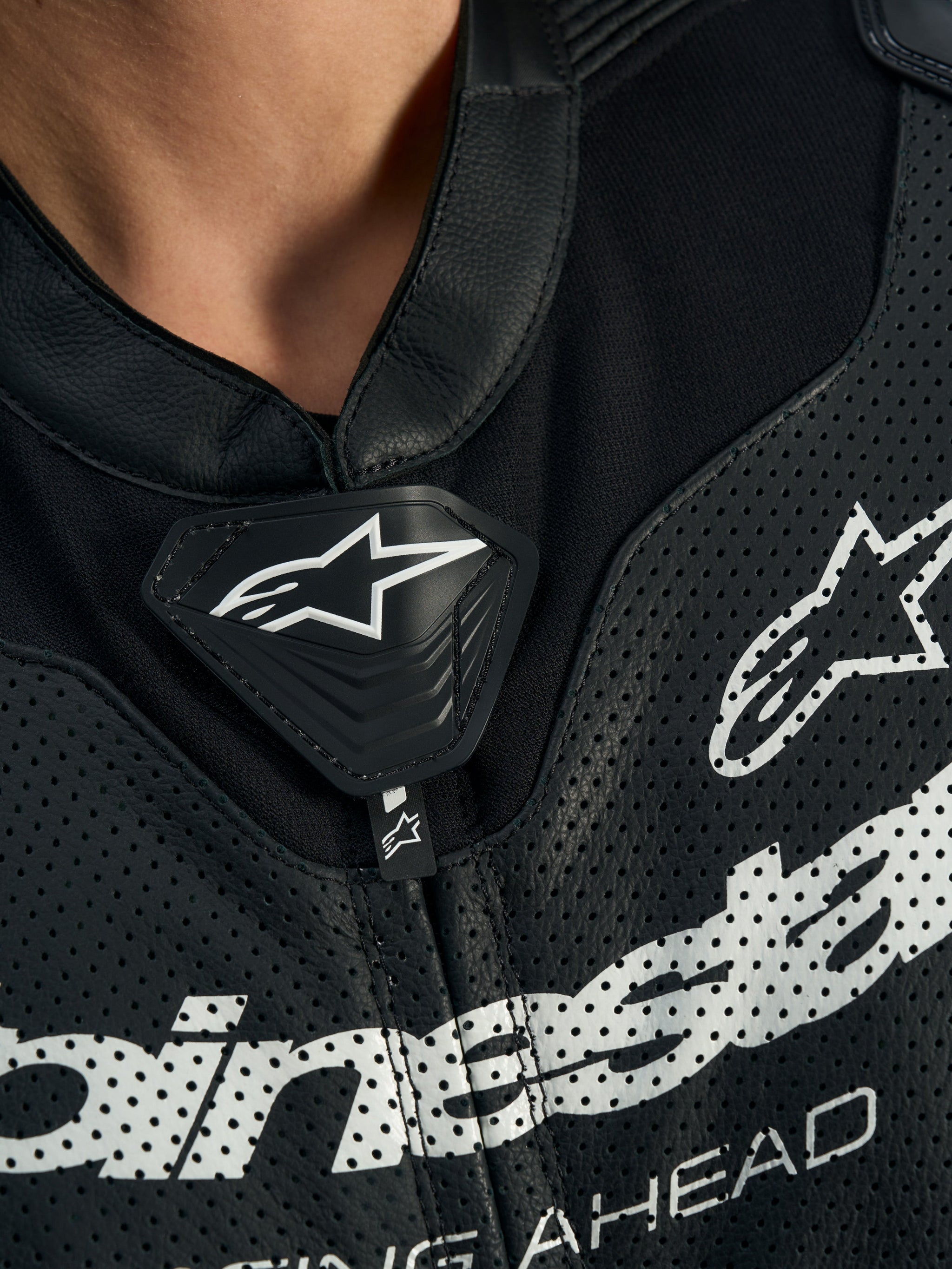Alpinestars Stella GP-R7 Leather Suit, fato de motociclismo de 1 peça para mulher, preto, pormenor aproximado do peito e da gola, com cabedal perfurado, logótipo central de escudo com a estrela da Alpinestars e grafismos brancos de grandes dimensões