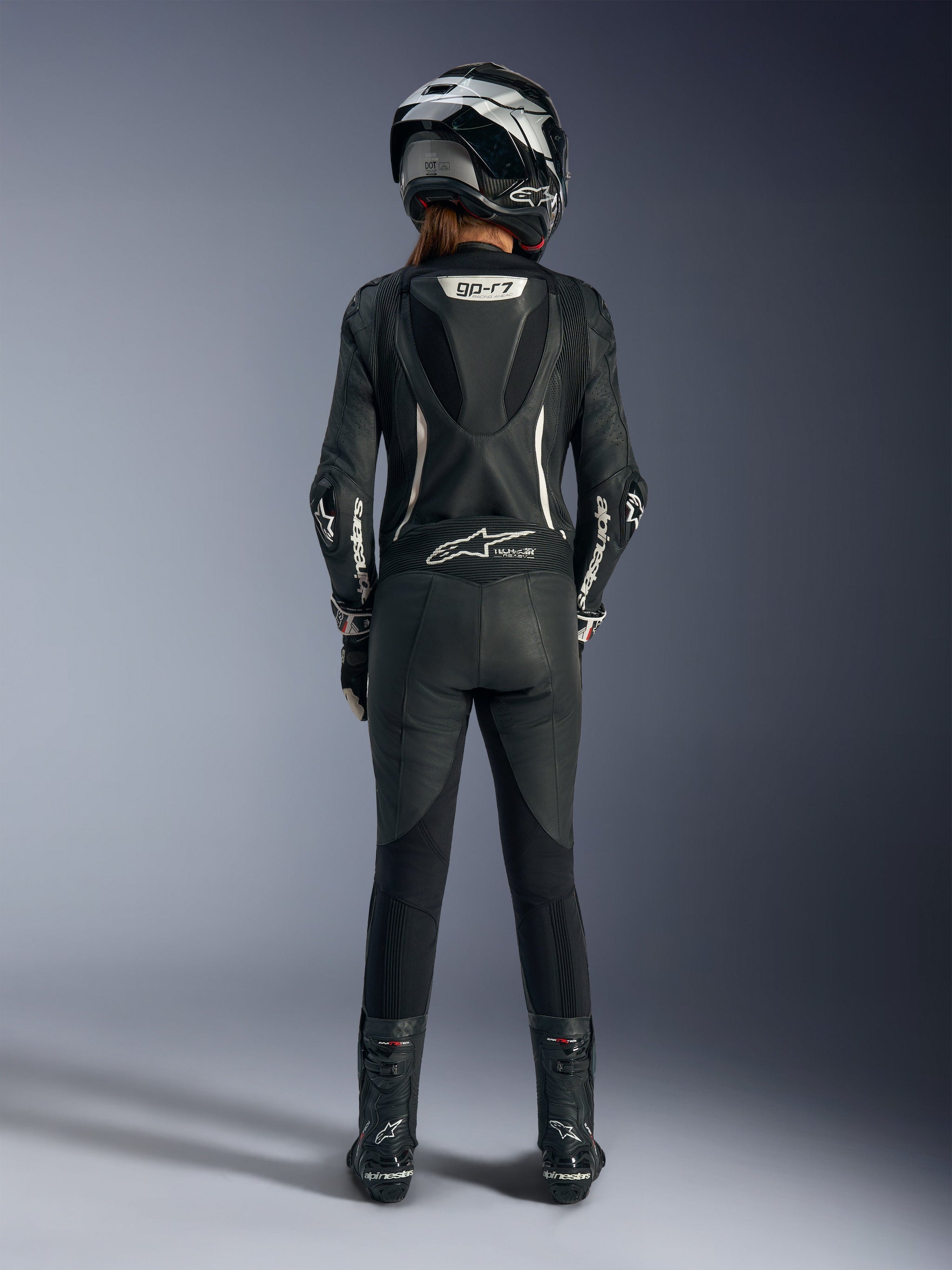 Piloto feminina a usar o fato de cabedal de uma peça Alpinestars Stella GP-R7 em preto, vista traseira, com detalhes em branco, corcova aerodinâmica com a marca 'GP-R7', combinado com um capacete de motociclismo prateado e preto e botas de corrida pretas.