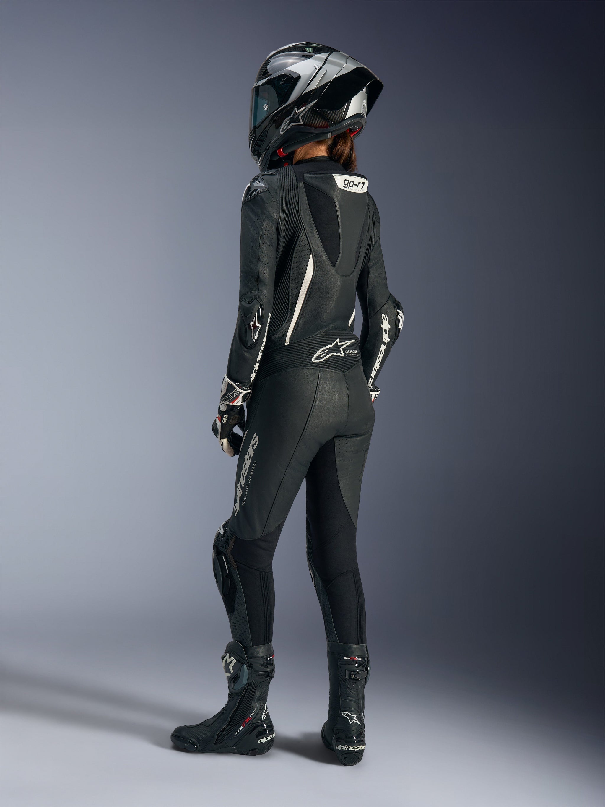 Piloto feminina vista de uma perspetiva traseira de três quartos a usar o Fato de Cabedal de Uma Peça Alpinestars Stella GP-R7 em preto. O fato apresenta logótipos brancos, uma corcova aerodinâmica proeminente com o texto 'GP-R7' e secções de cabedal perfurado. O conjunto é complementado por um capacete de corrida integral em preto e prateado a condizer, luvas pretas e brancas e botas pretas para um equipamento de pista profissional completo.