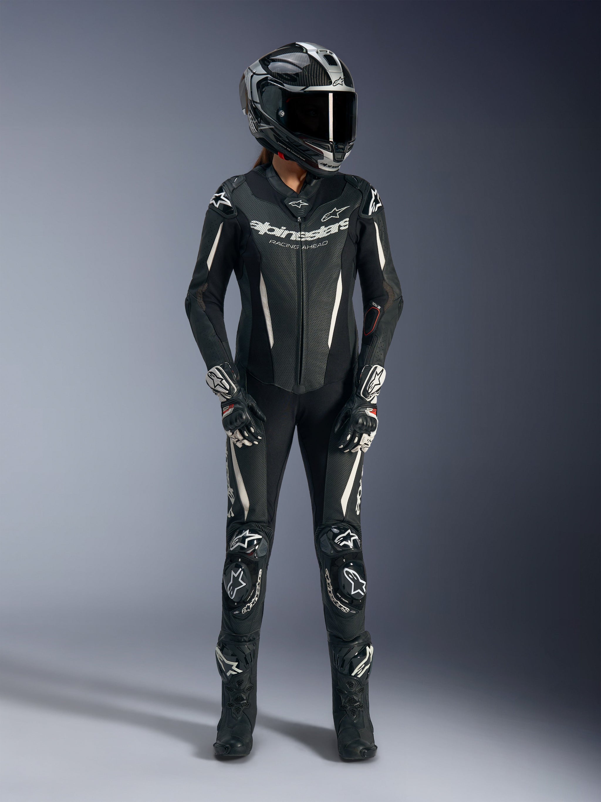 Piloto feminina a usar o fato de cabedal de uma peça Alpinestars Stella GP-R7 em preto, equipamento completo de corrida de motociclismo incluindo capacete Supertech R10, luvas pretas e brancas e botas de corrida, com pormenores em branco, painéis de cabedal perfurado e bossa aerodinâmica para desempenho profissional em pista.