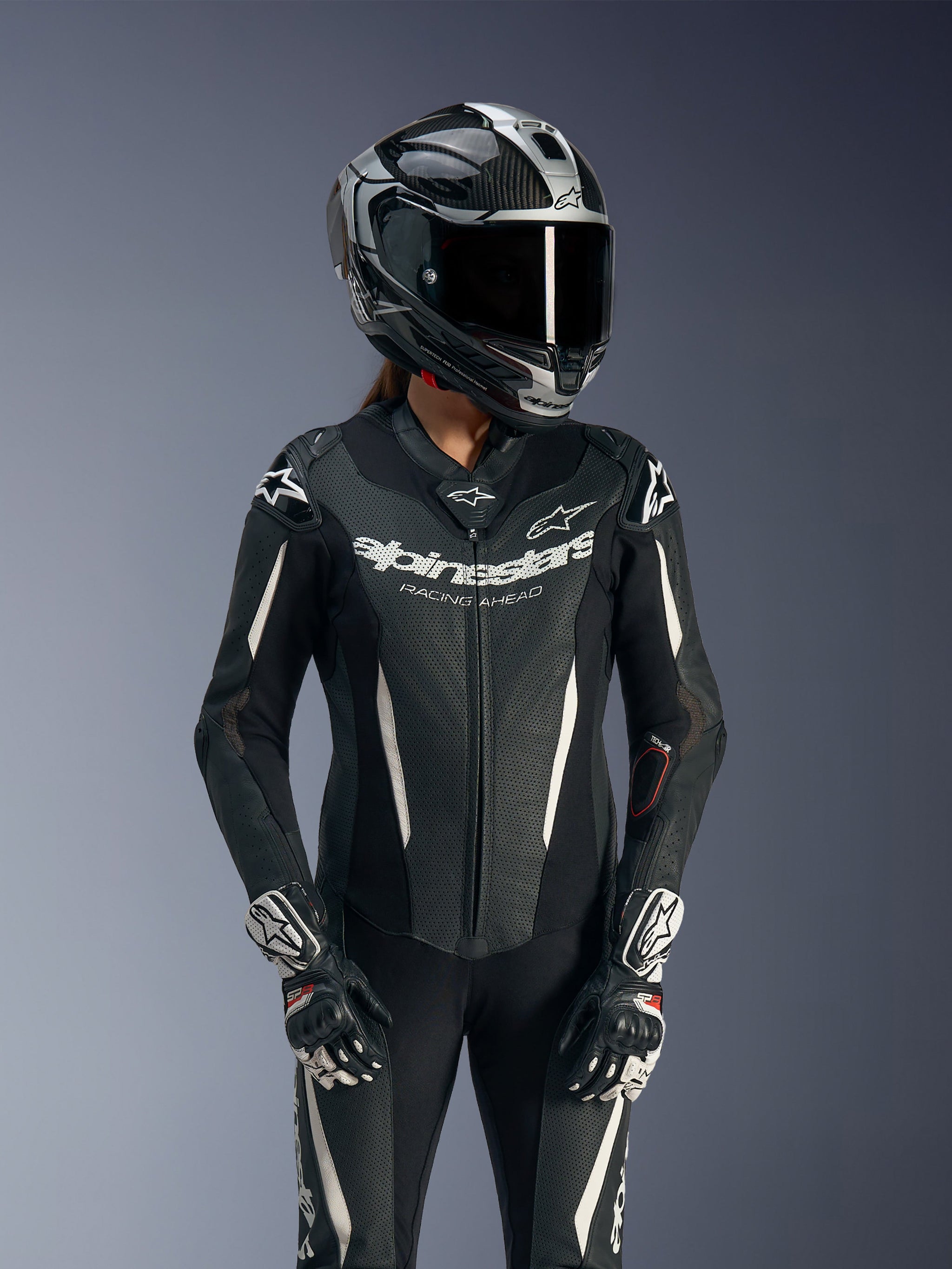 Piloto feminina com Fato de Cabedal de Uma Peça Alpinestars Stella GP-R7, preto com detalhes brancos, usado com capacete de corrida e luvas, com painéis de cabedal perfurado e logótipo Alpinestars 'Racing Ahead' no peito, equipamento profissional de pista.