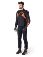 Casaco Alpinestars T-Jaws V4 Waterproof, preto e vermelho fluo, usado por um modelo masculino com calças, luvas e botas pretas, apresentando detalhes em laranja vibrante e o logótipo Alpinestars, design desportivo para motociclismo