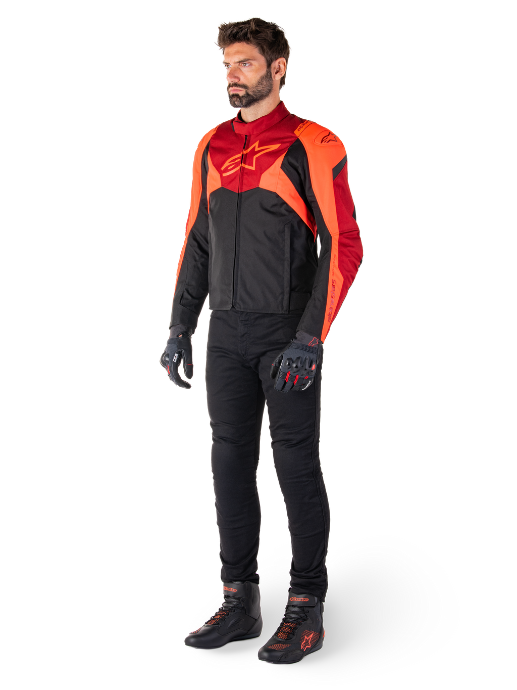 Piloto masculino a usar o Casaco Impermeável Alpinestars T-Jaws V4 em preto, vermelho fluo e vermelho escuro, combinado com calças de motociclismo pretas, luvas e botas, apresentando um design têxtil de ajuste desportivo com logótipo proeminente no peito e uma silhueta agressiva.