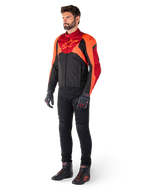 Piloto masculino a usar o Casaco Impermeável Alpinestars T-Jaws V4 em preto, vermelho fluo e vermelho escuro, combinado com calças de motociclismo pretas, luvas e botas, apresentando um design têxtil de ajuste desportivo com logótipo proeminente no peito e uma silhueta agressiva.