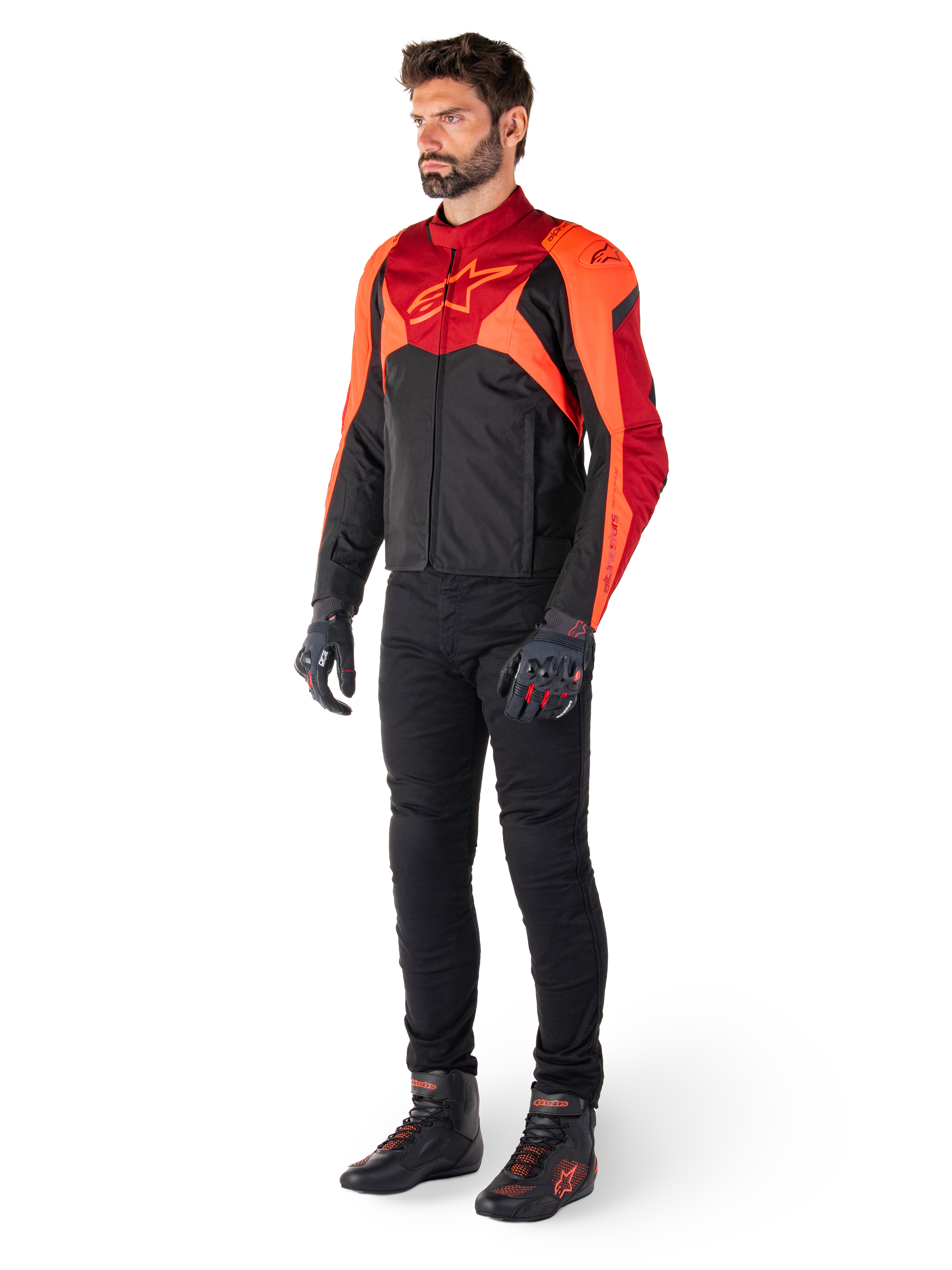 Piloto masculino a usar o Casaco Impermeável Alpinestars T-Jaws V4 em preto, vermelho fluo e vermelho escuro, combinado com calças de motociclismo pretas, luvas e botas, apresentando um design têxtil de ajuste desportivo com logótipo proeminente no peito e uma silhueta agressiva.
