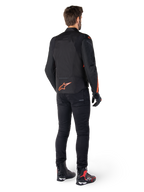 Casaco Impermeável Alpinestars T-Jaws V4, preto e vermelho fluo, vista traseira usado por um piloto masculino, com logótipo vermelho fluo vibrante nas costas, calças de condução pretas e botas técnicas, vestuário de motociclismo desportivo.