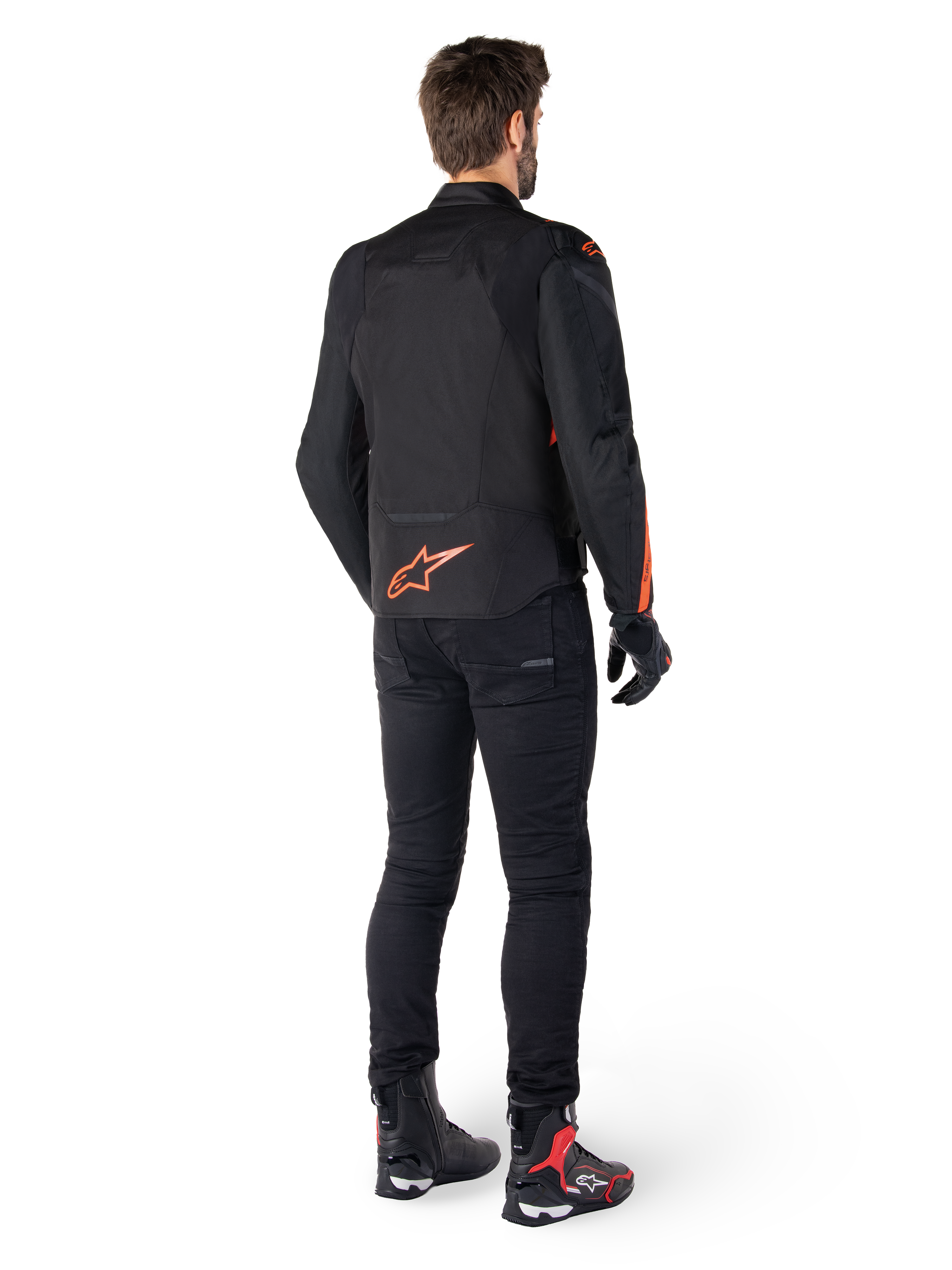 Casaco Impermeável Alpinestars T-Jaws V4, preto e vermelho fluo, vista traseira usado por um piloto masculino, com logótipo vermelho fluo vibrante nas costas, calças de condução pretas e botas técnicas, vestuário de motociclismo desportivo.