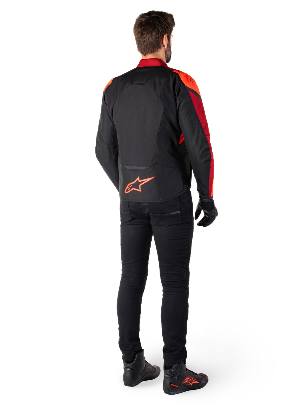 Casaco Impermeável Alpinestars T-Jaws V4 em Preto Vermelho Fluo Vermelho Escuro, vista traseira de um piloto a usar o casaco têxtil com calças pretas e botas de condução, apresentando um logótipo Alpinestars vermelho de destaque na parte inferior das costas.