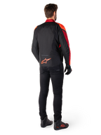 Casaco Impermeável Alpinestars T-Jaws V4 em Preto Vermelho Fluo Vermelho Escuro, vista traseira de um piloto a usar o casaco têxtil com calças pretas e botas de condução, apresentando um logótipo Alpinestars vermelho de destaque na parte inferior das costas.