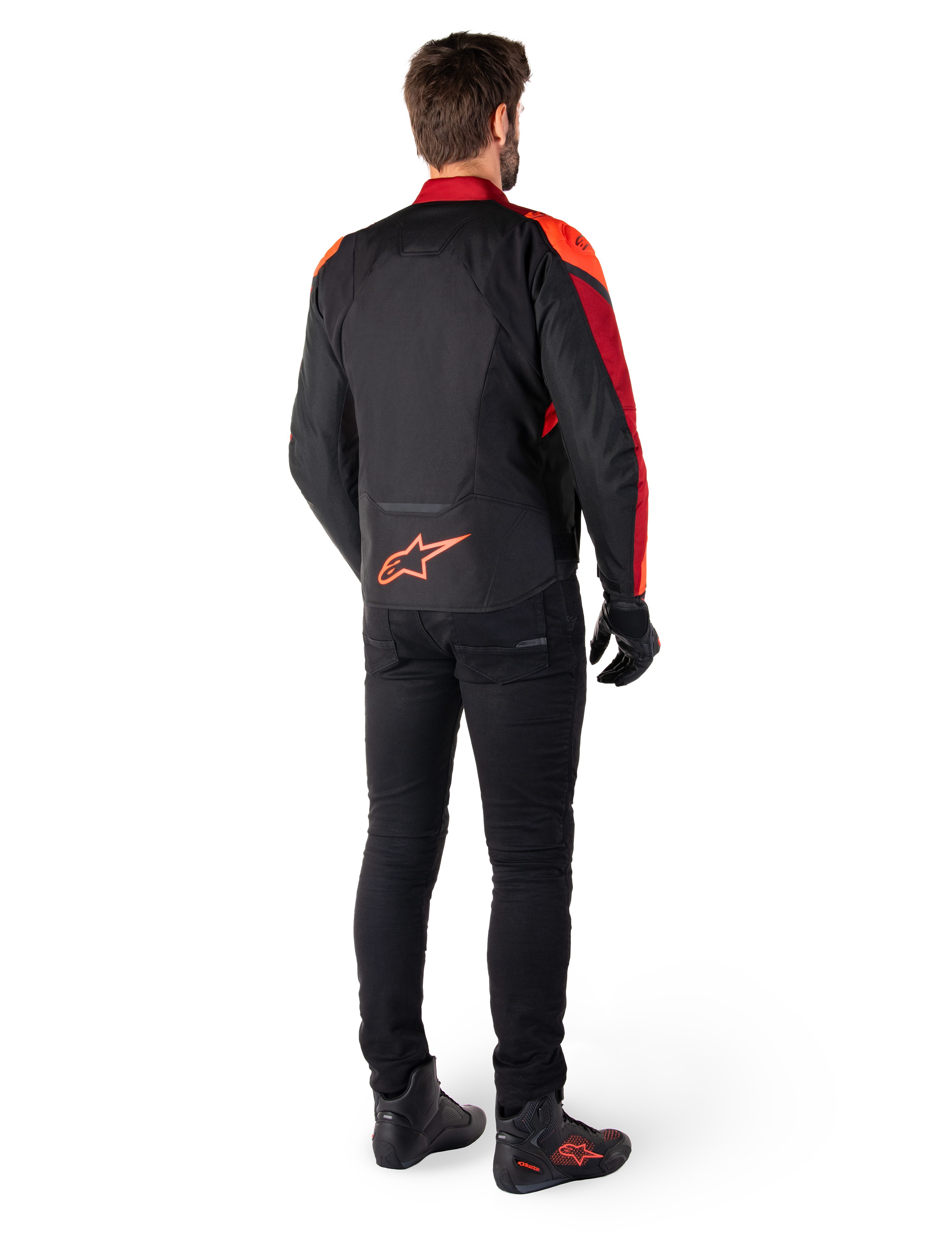 Casaco Impermeável Alpinestars T-Jaws V4 em Preto Vermelho Fluo Vermelho Escuro, vista traseira de um piloto a usar o casaco têxtil com calças pretas e botas de condução, apresentando um logótipo Alpinestars vermelho de destaque na parte inferior das costas.