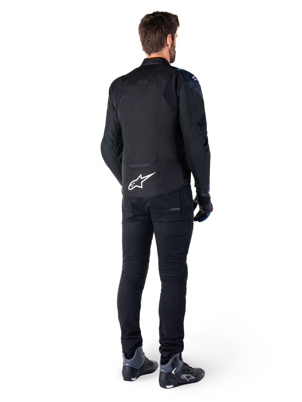 Vista traseira de um homem a usar o casaco impermeável Alpinestars T-Jaws V4 em preto, azul escuro e azul Blithe, com um logótipo Astar branco proeminente na parte inferior das costas, combinado com calças pretas e sapatos de motociclismo.