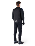 Vista traseira de um homem a usar o casaco impermeável Alpinestars T-Jaws V4 em preto, azul escuro e azul Blithe, com um logótipo Astar branco proeminente na parte inferior das costas, combinado com calças pretas e sapatos de motociclismo.