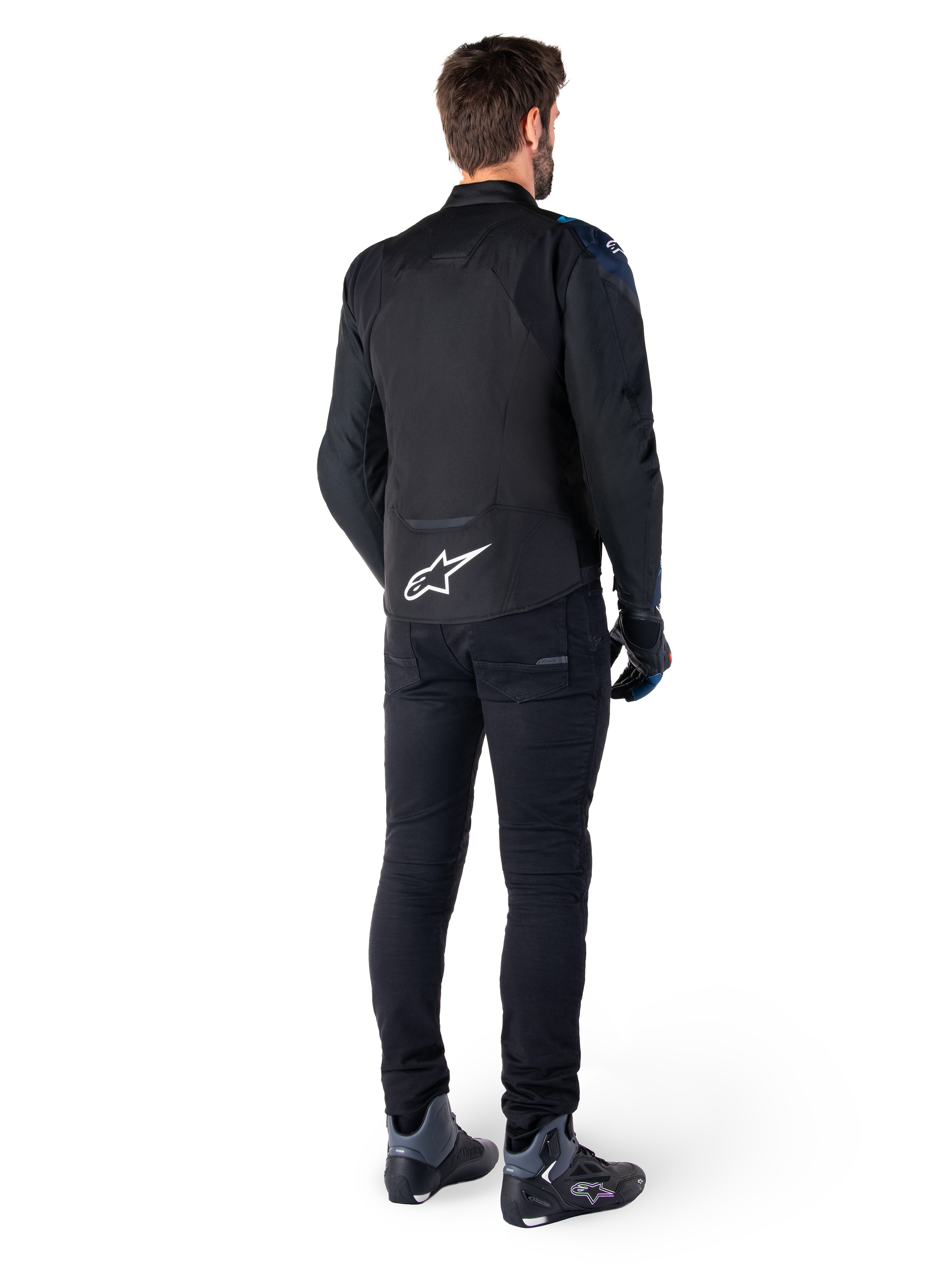Vista traseira de um homem a usar o casaco impermeável Alpinestars T-Jaws V4 em preto, azul escuro e azul Blithe, com um logótipo Astar branco proeminente na parte inferior das costas, combinado com calças pretas e sapatos de motociclismo.