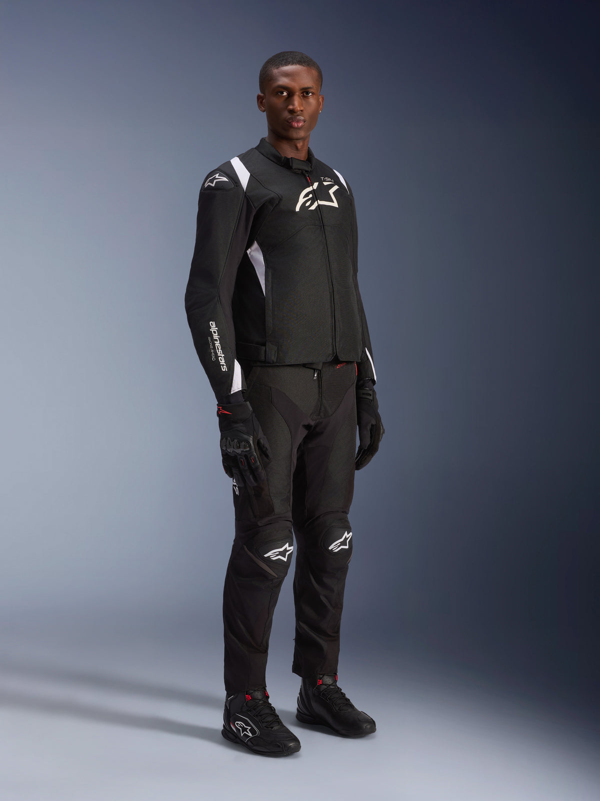 Modelo masculino a usar o Casaco Impermeável Alpinestars T-SP 1 V2 em preto e branco, acompanhado por calças, luvas e botas de motociclismo pretas. Fotografia de estúdio de corpo inteiro que destaca o casaco têxtil desportivo com logótipos brancos e uma silhueta ajustada em 3D.