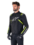 Modelo masculino a usar o casaco Alpinestars T-SPS V2 Waterproof, preto com detalhes em branco e amarelo fluo, com o logótipo Astars branco no peito e riscas amarelo fluorescente nos ombros e mangas, combinado com calças pretas e luvas de motociclismo, casaco de motociclismo têxtil com corte desportivo.