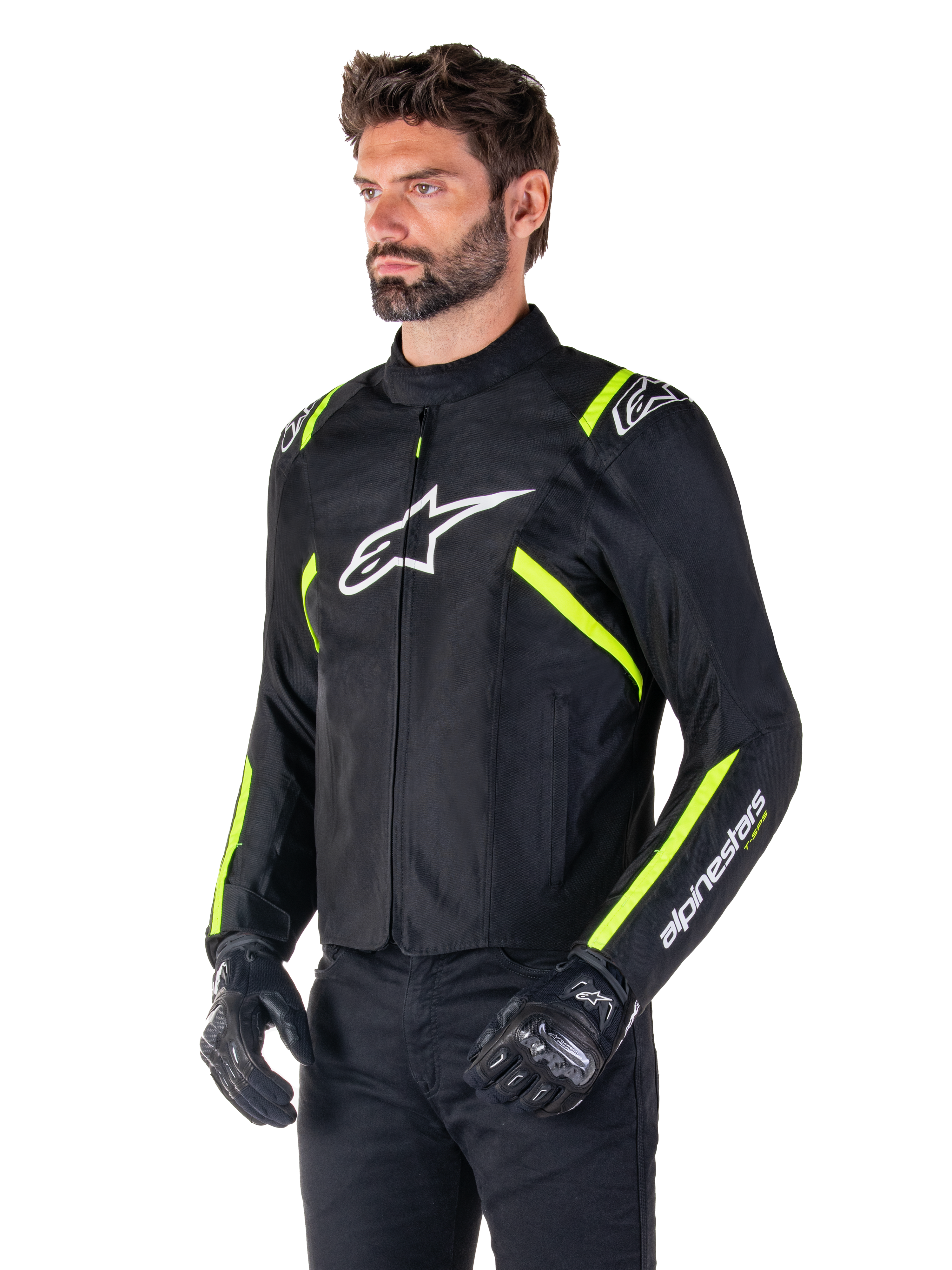 Modelo masculino a usar o casaco Alpinestars T-SPS V2 Waterproof, preto com detalhes em branco e amarelo fluo, com o logótipo Astars branco no peito e riscas amarelo fluorescente nos ombros e mangas, combinado com calças pretas e luvas de motociclismo, casaco de motociclismo têxtil com corte desportivo.