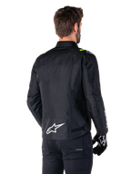 Alpinestars T-SPS V2 Waterproof Jacket, Preto Branco Amarelo Fluo, vista traseira de modelo masculino a usar o casaco de motociclismo têxtil, com logótipo branco na parte inferior das costas e detalhes em amarelo fluo nos ombros, combinado com calças e luvas pretas.