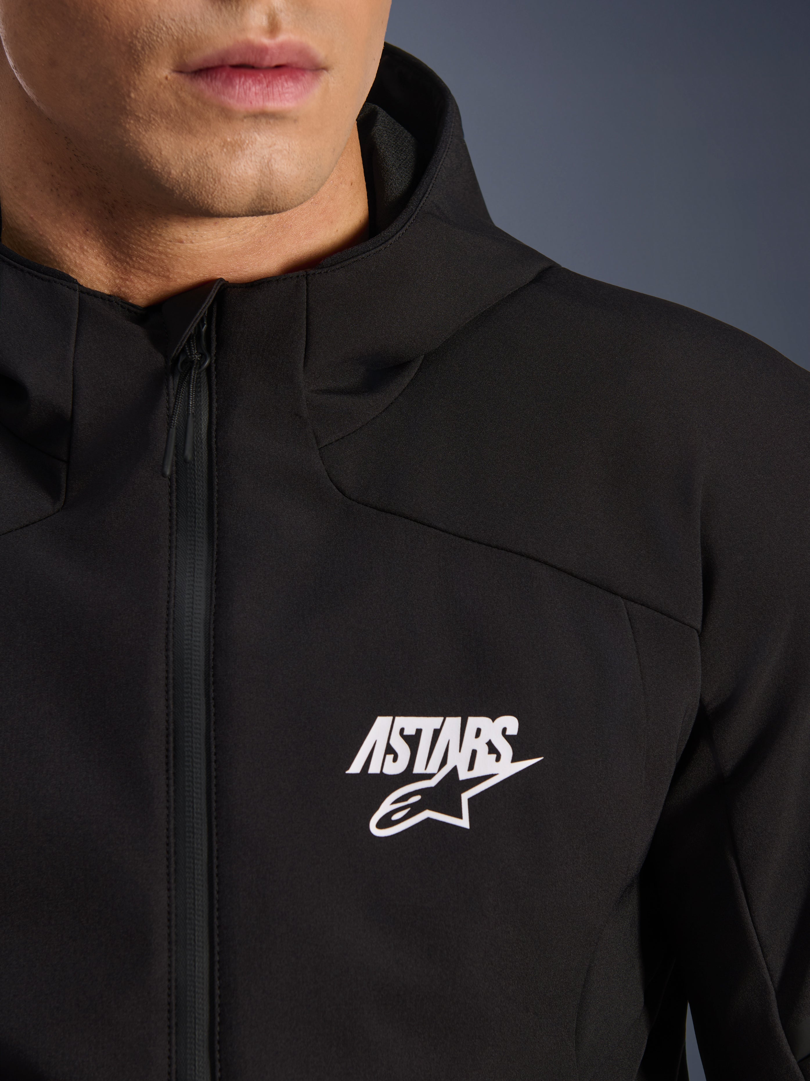 Alpinestars Aeroshell Waterproof Jacket, black motorcycle softshell jacket, close-up view of the chest featuring a white ASTARS star logo and waterproof front zipper, integrated hood, modern sport-styled design