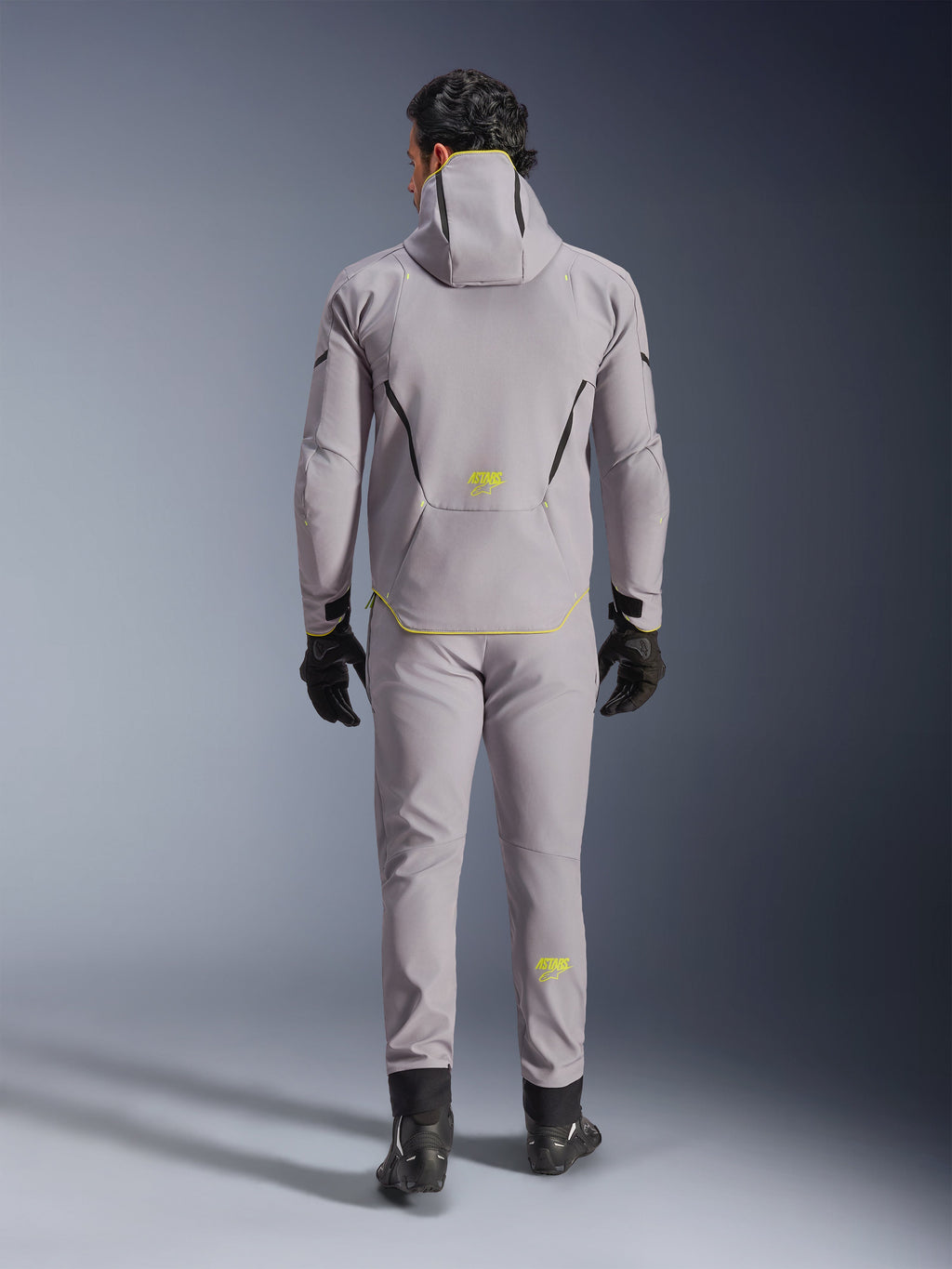 Casaco Alpinestars Aeroshell WP em cinzento e amarelo fluo, vista traseira completa, usado com calças cinzentas a condizer, luvas pretas e sapatos de condução pretos. Apresenta um design softshell com capuz, logótipo 'ASTARS' e detalhes em amarelo fluo, concebido para performance moderna de motociclismo em estrada.
