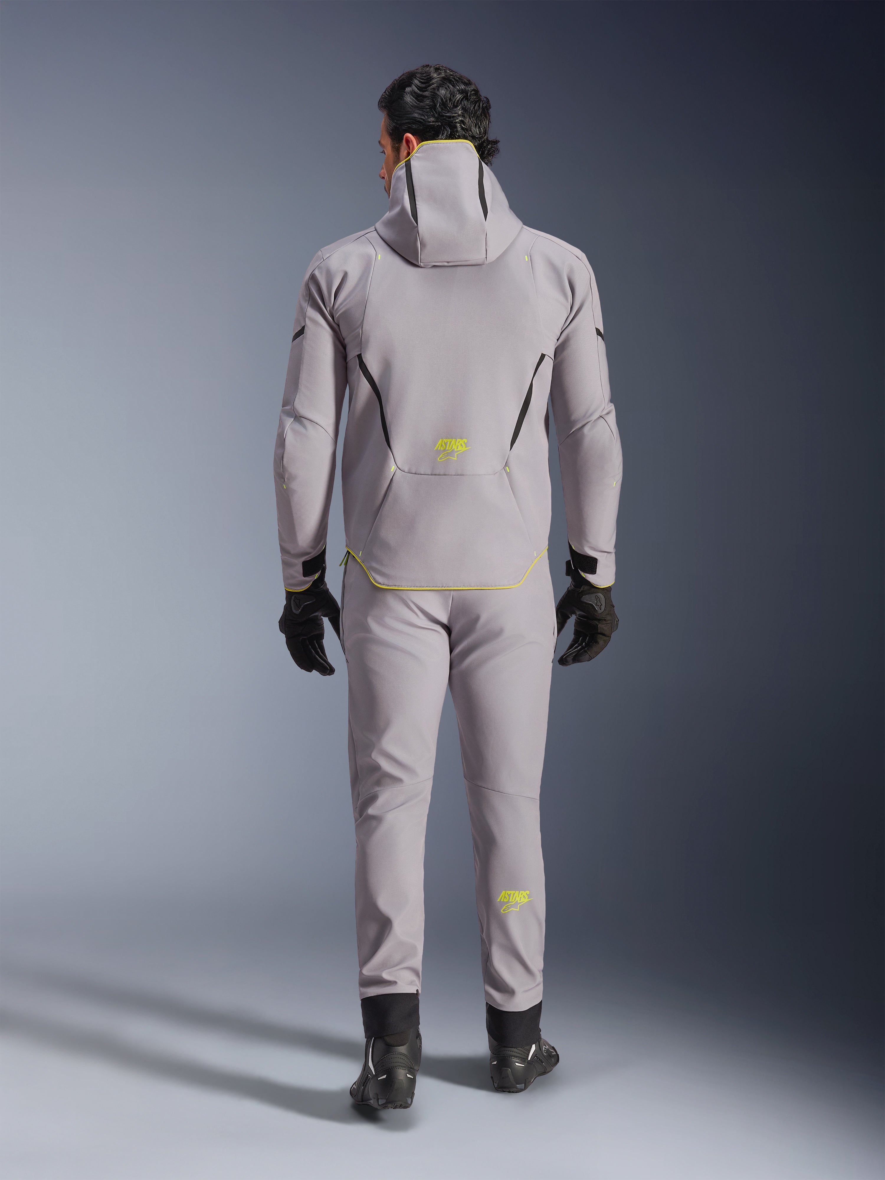 Casaco Alpinestars Aeroshell WP em cinzento e amarelo fluo, vista traseira completa, usado com calças cinzentas a condizer, luvas pretas e sapatos de condução pretos. Apresenta um design softshell com capuz, logótipo 'ASTARS' e detalhes em amarelo fluo, concebido para performance moderna de motociclismo em estrada.