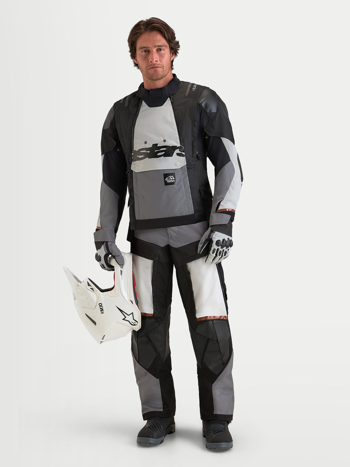 Alpinestars Harok V 2 Drystar casaco de moto para homem, todo-o-terreno, cinzento-escuro, cinzento-gelo e preto, construção têxtil impermeável com proteção certificada CE, vestido por modelo masculino a segurar um capacete branco