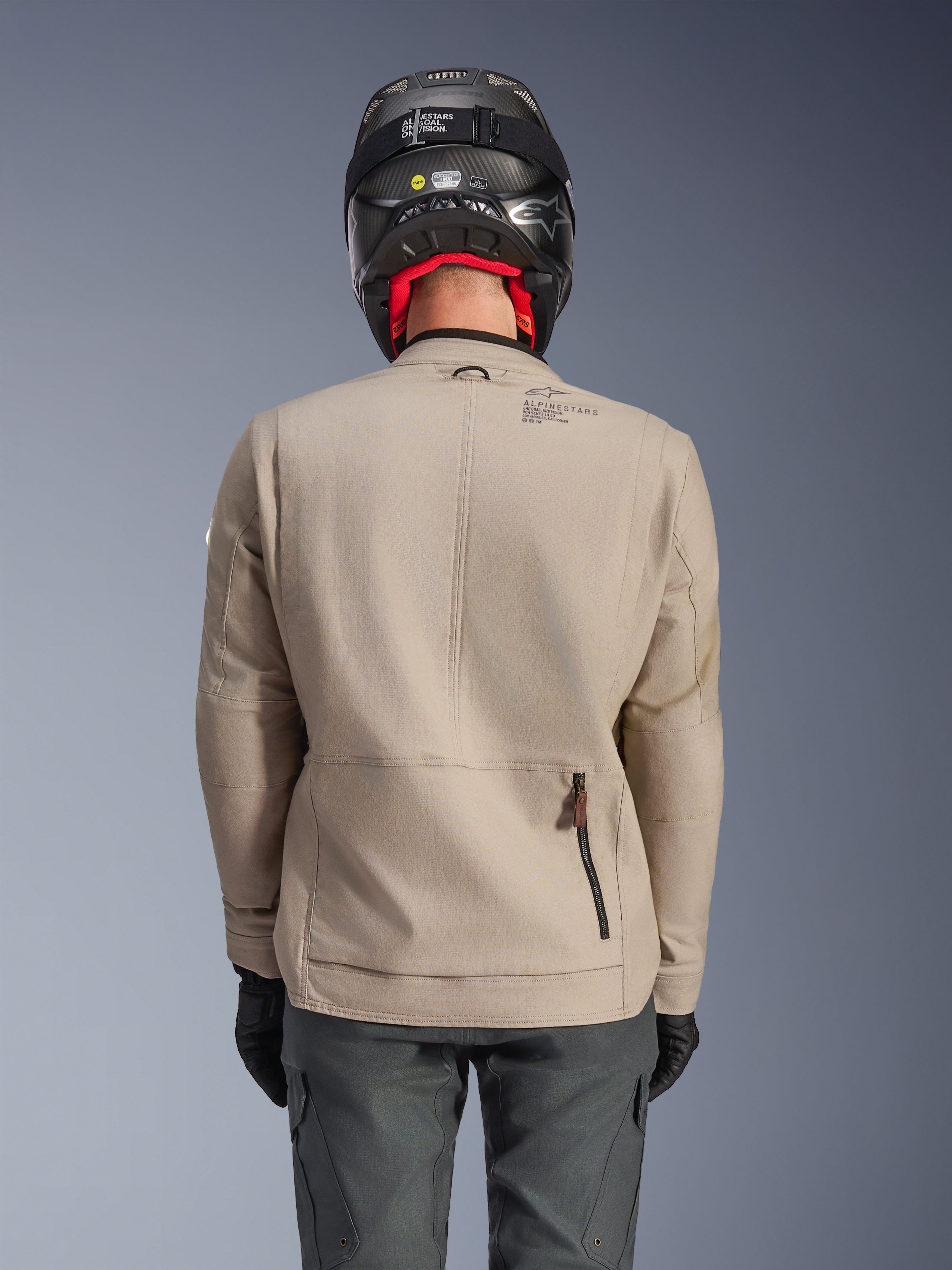 Casaco Alpinestars Borrego Drystar®, casaco de motociclismo de aventura, cinzento quente, vista traseira mostrando o fecho do compartimento traseiro e a marca Alpinestars, usado com um capacete preto e calças de motociclismo cinzento escuro