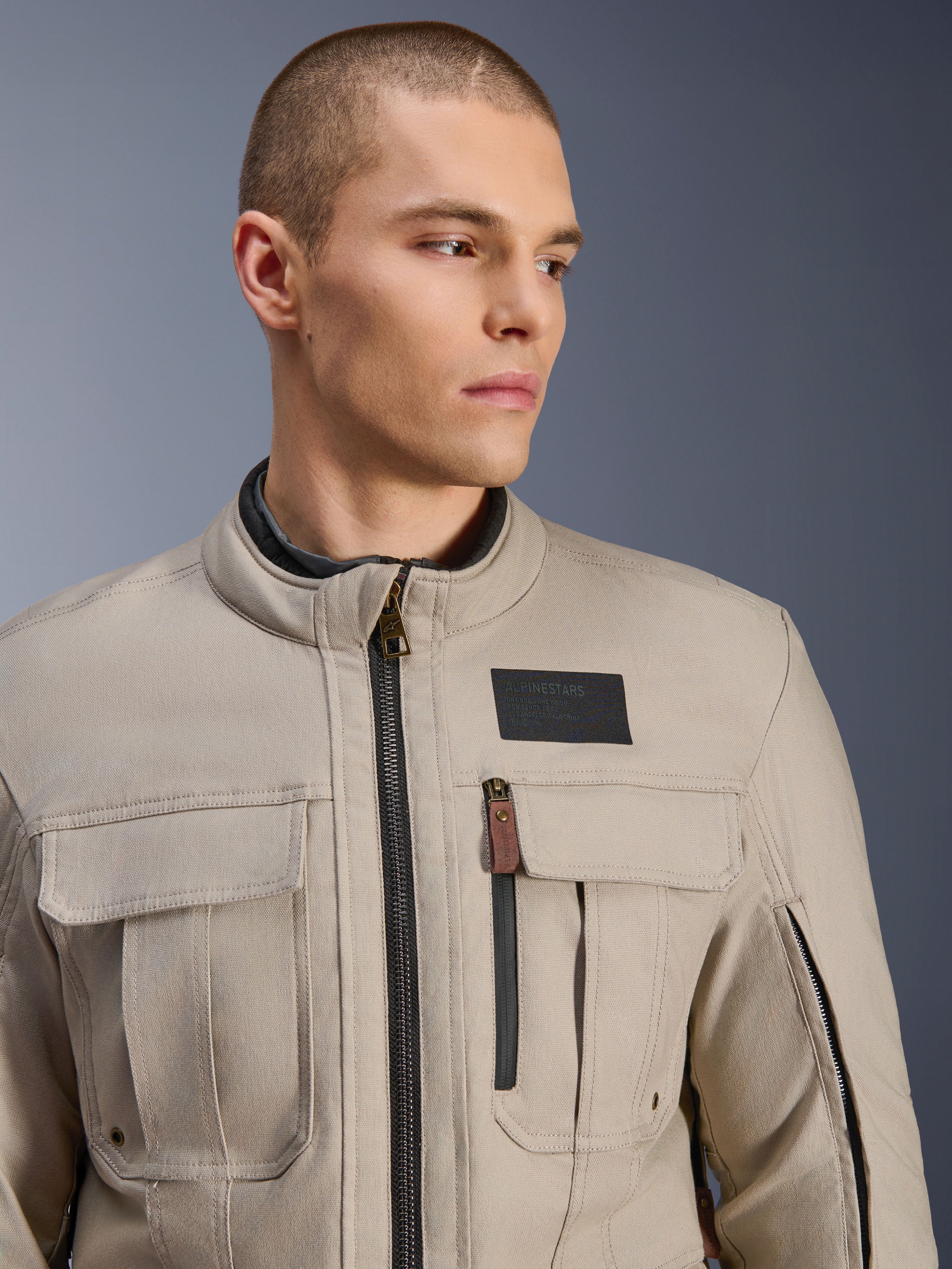 Modelo masculino a usar o Casaco Alpinestars Borrego Drystar® em Warm Gray, casaco de motociclismo de aventura com visual clássico de lona encerada, bolsos de carga funcionais no peito e um logótipo Alpinestars preto.