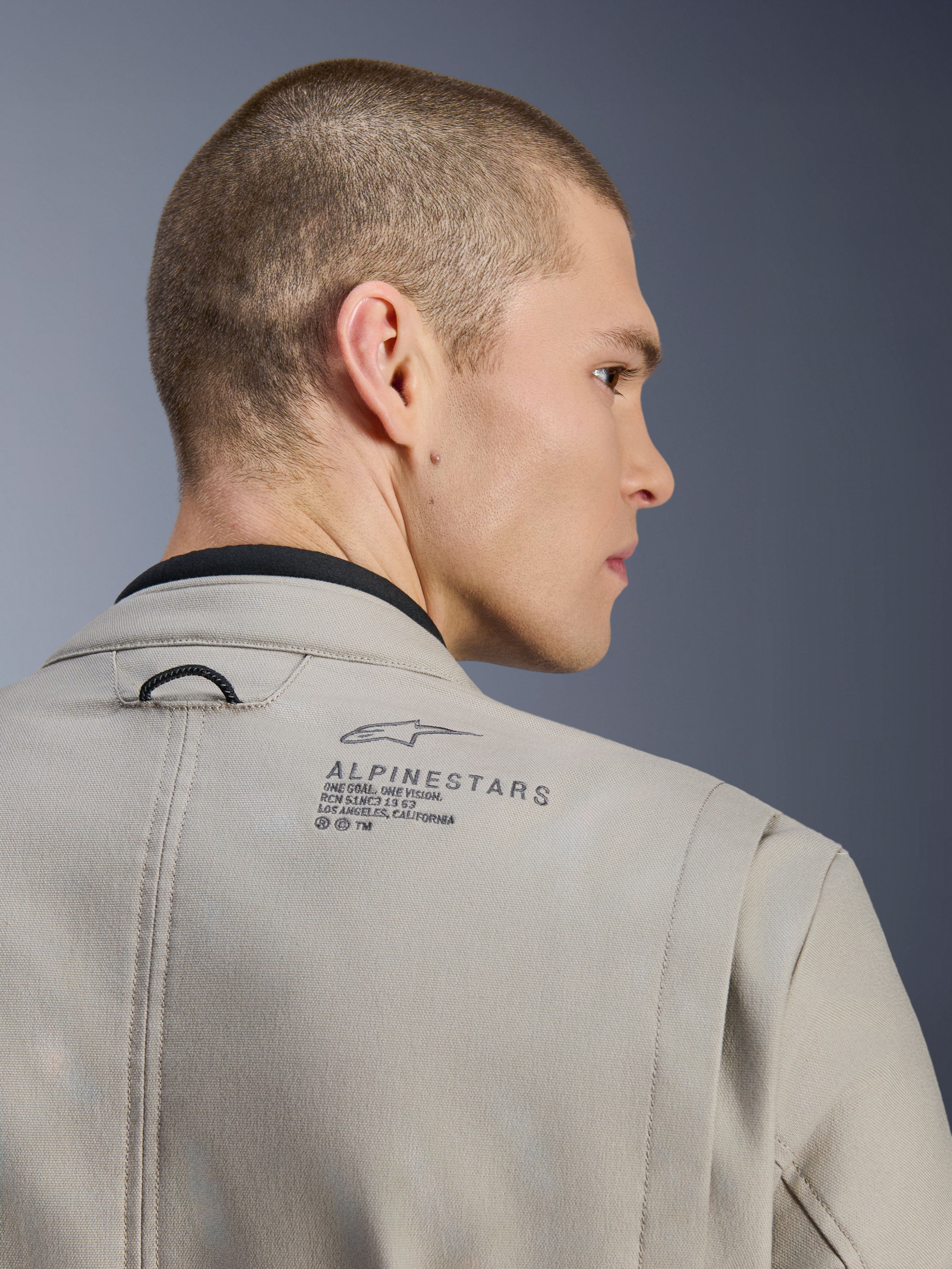 Casaco Alpinestars Borrego Drystar® em Warm Gray, detalhe do ombro traseiro, material de lona encerada texturizada, com o logótipo Alpinestars preto e texto heritage, usado por um modelo, casaco de motociclismo de aventura