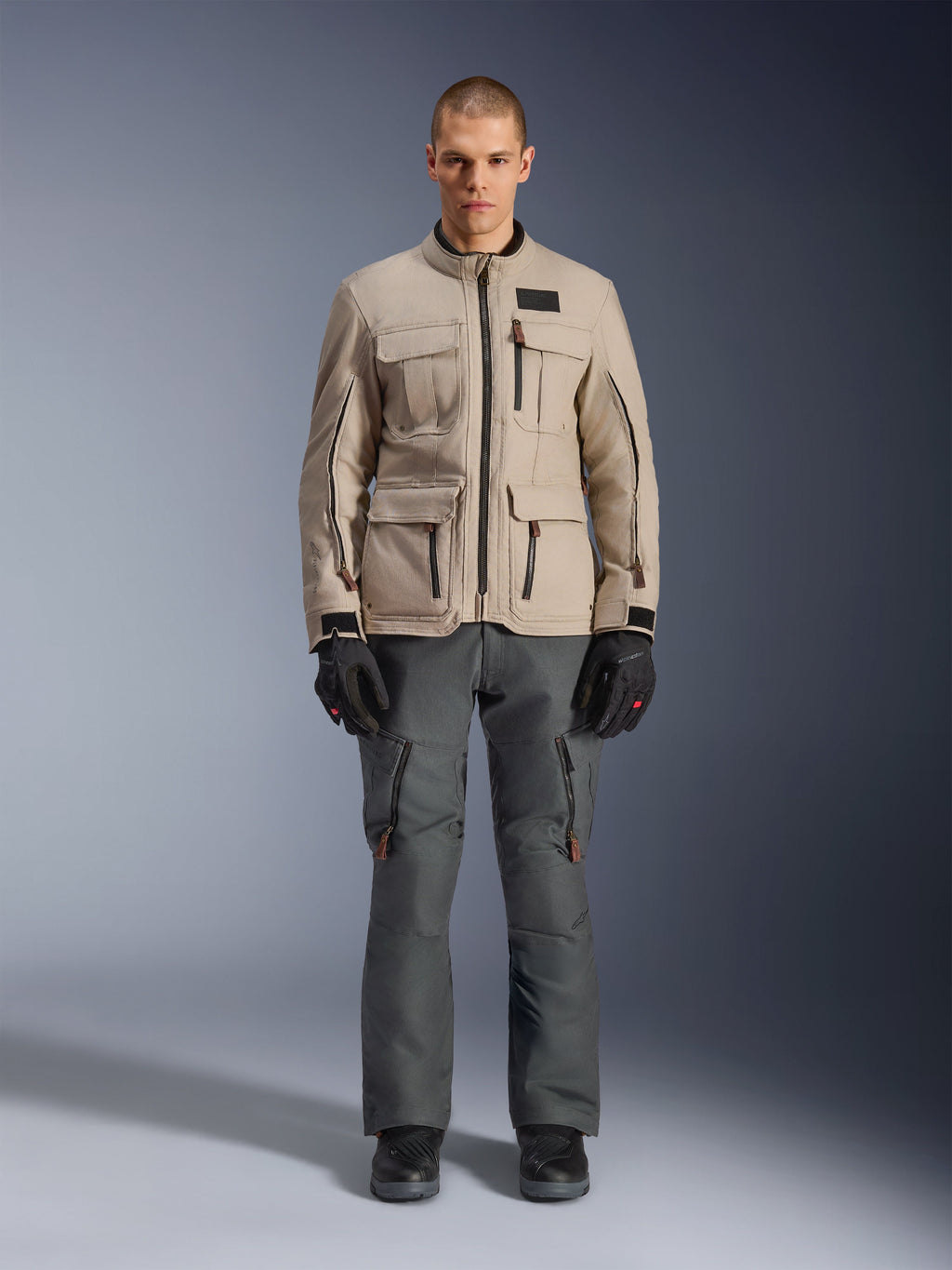 Casaco Alpinestars Borrego Drystar®, casaco de motociclismo de aventura, cinzento (Warm Gray), usado por um modelo com calças de motociclismo cinzento-escuro e luvas pretas, com vários bolsos de carga e uma construção têxtil durável com um aspeto clássico de lona encerada.